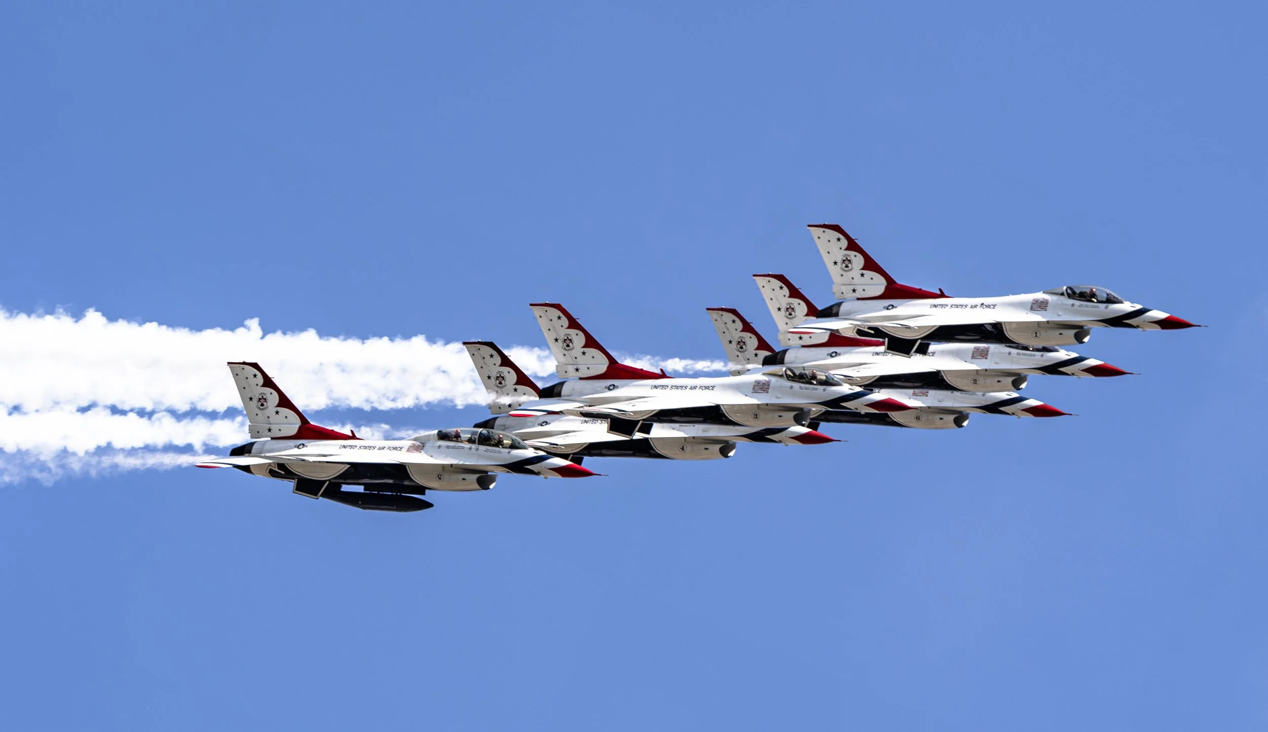 The Thunderbirds climb in tight formation, leaving bright white smoke trails that cut across the summer sky.