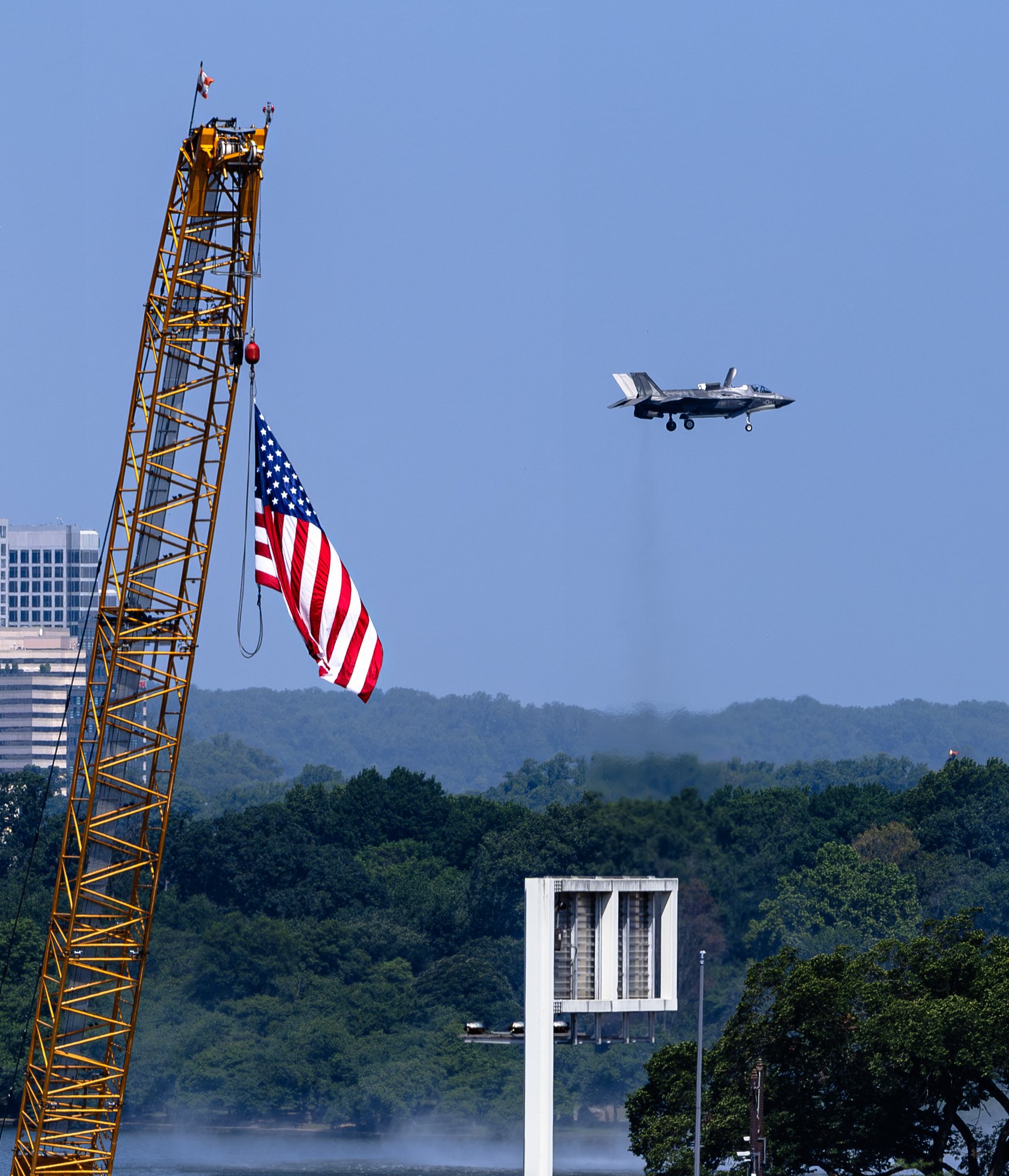 An F-35 Lightning II hovers next to a construction crane displaying a large American flag, symbolizing modern military aviation and American industry during the celebration.