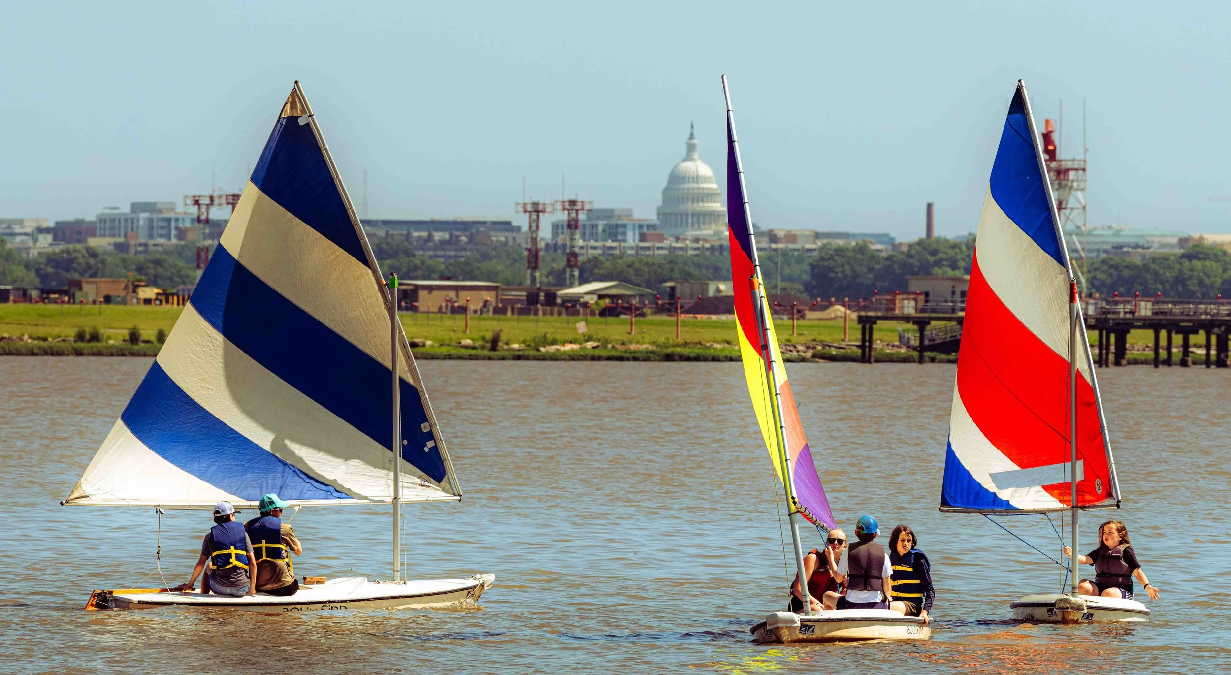 A sailor guides a dinghy beneath a multicolored sail with Washington landmarks visible across the river.