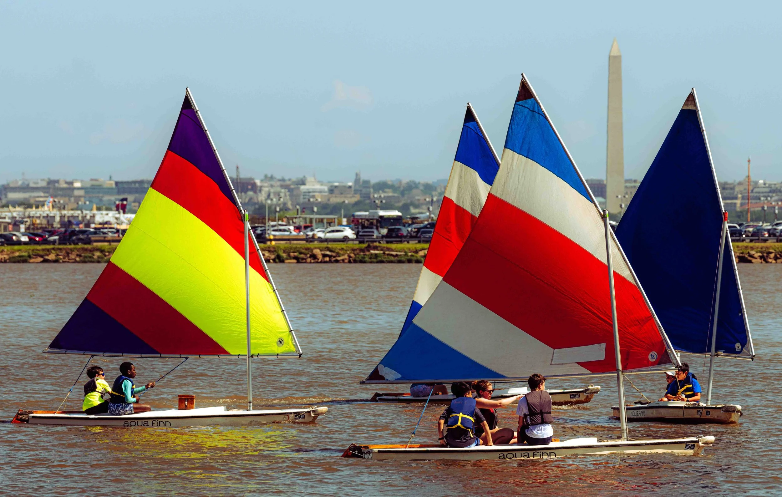 Sailing students navigate the river with views of Washington, D.C.'s iconic landmarks in the background.