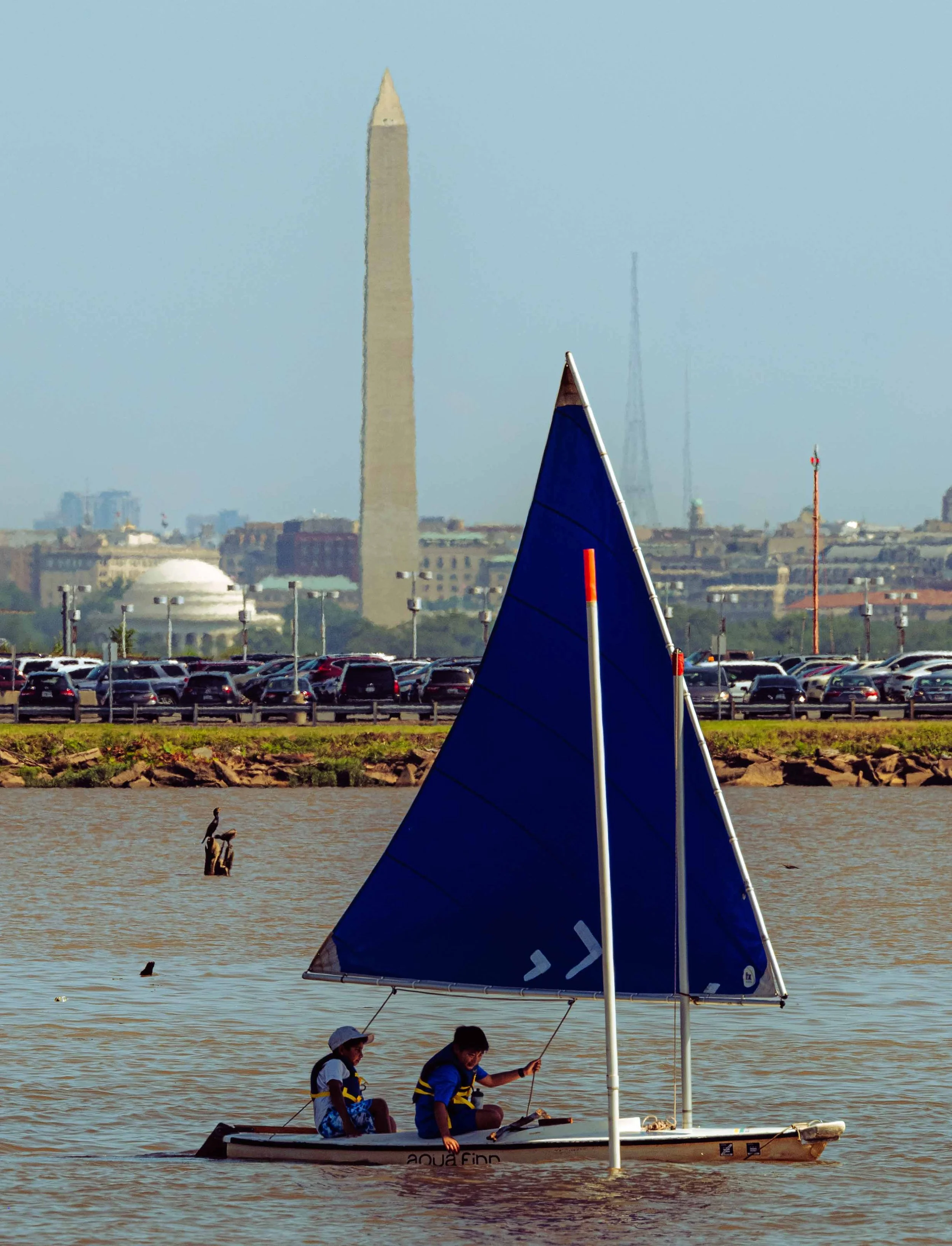 A student sailor navigates beneath a blue sail with the Washington Monument visible across the river.