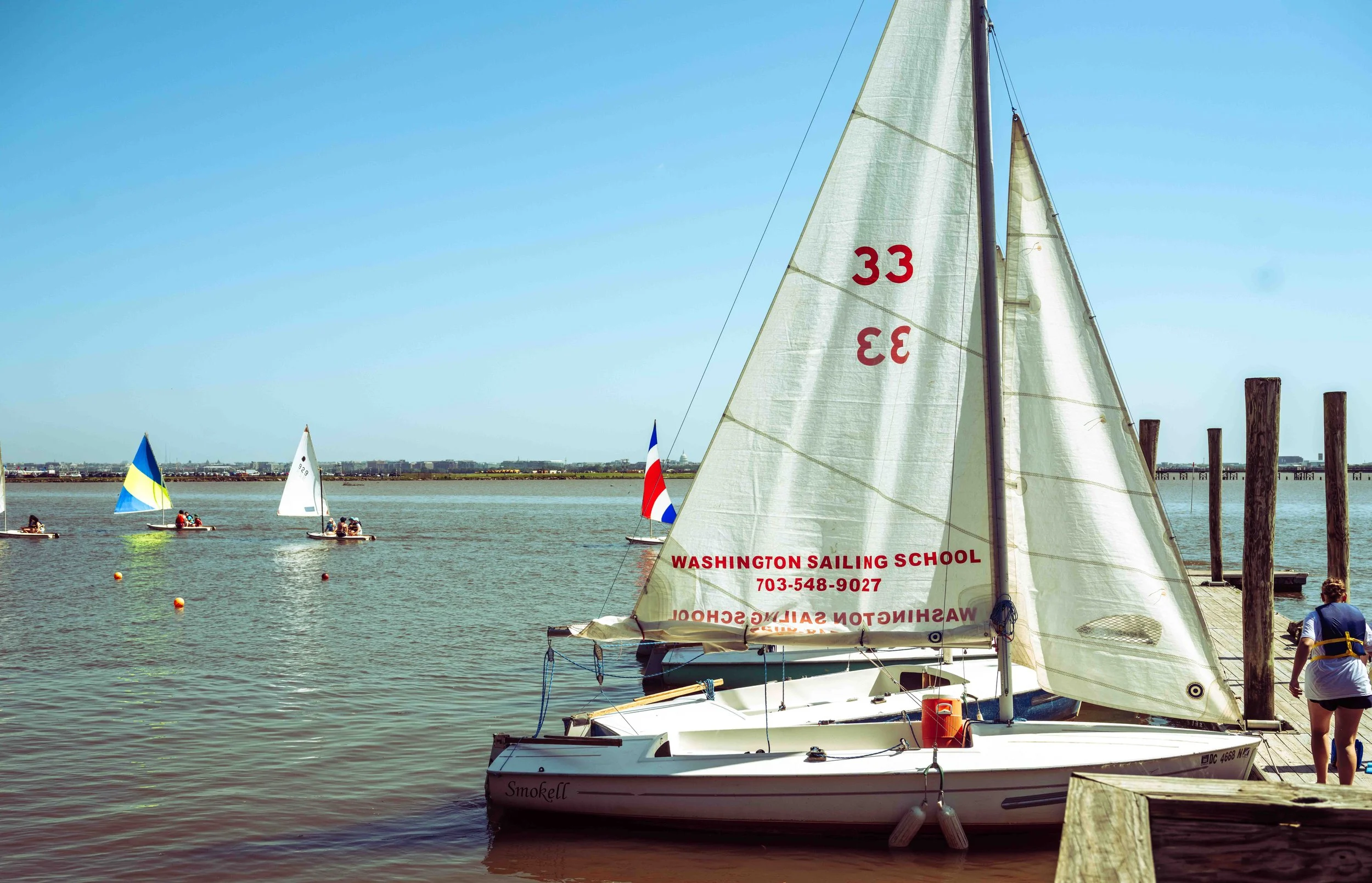 Brightly colored sails reflect on the water as young sailors gather near the dock.