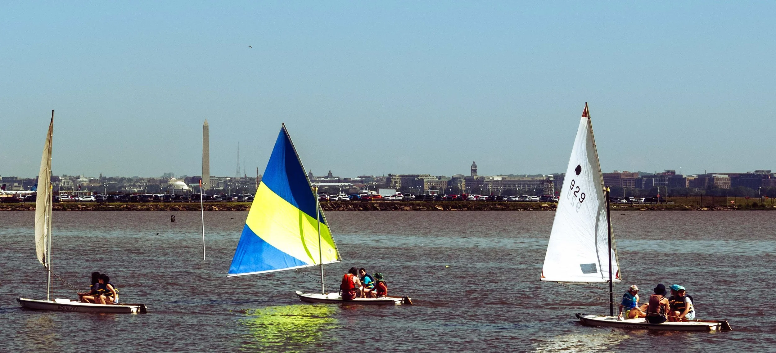 Student sailors navigate the Potomac River with the Washington Monument rising above the horizon.