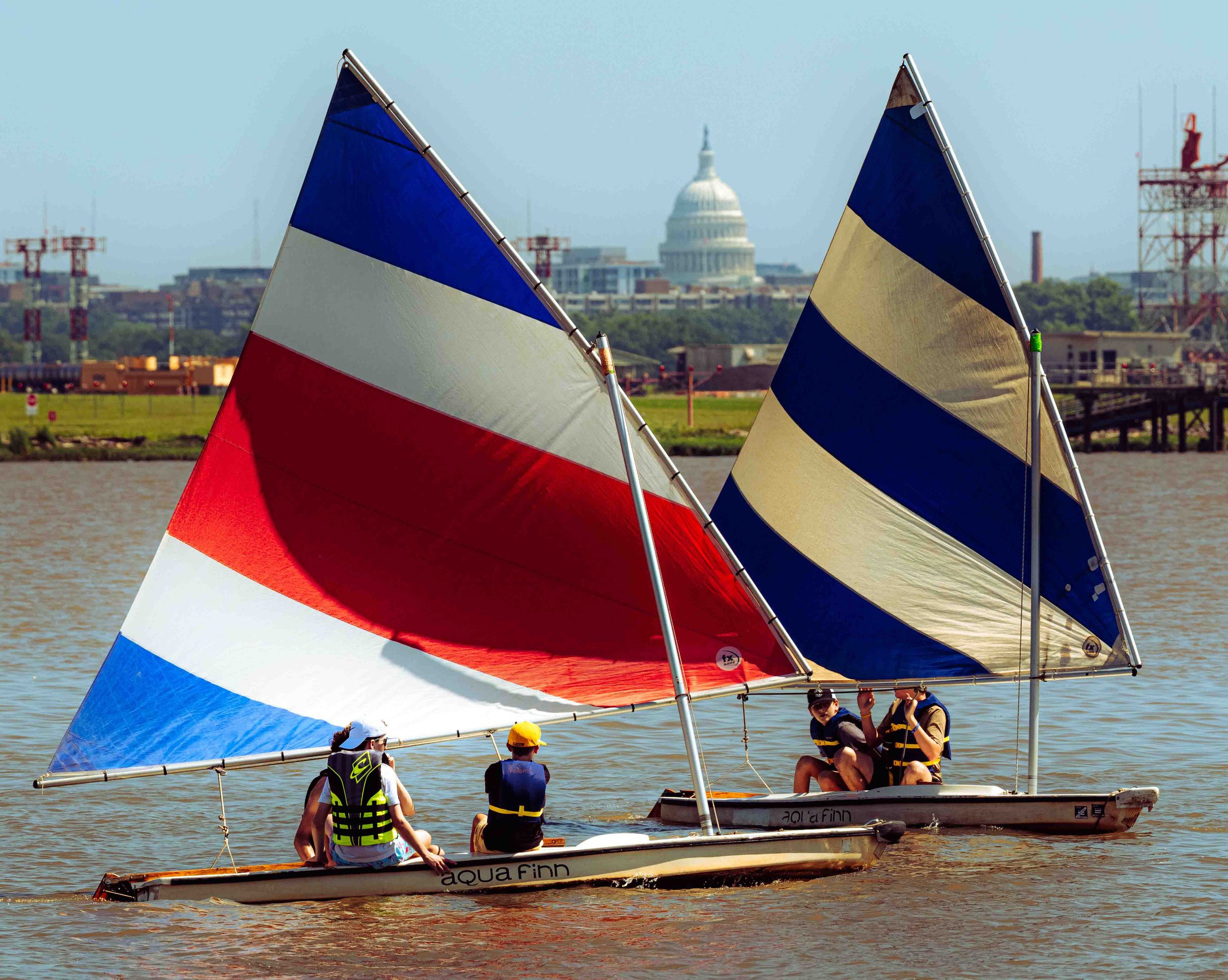 Young sailors race across the river under blue, red, and white sails near the Washington waterfront.