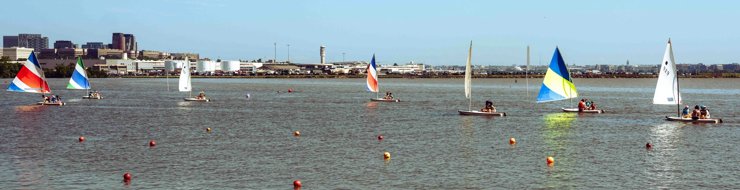 Colorful dinghies sail through a marked practice area during a youth sailing lesson.