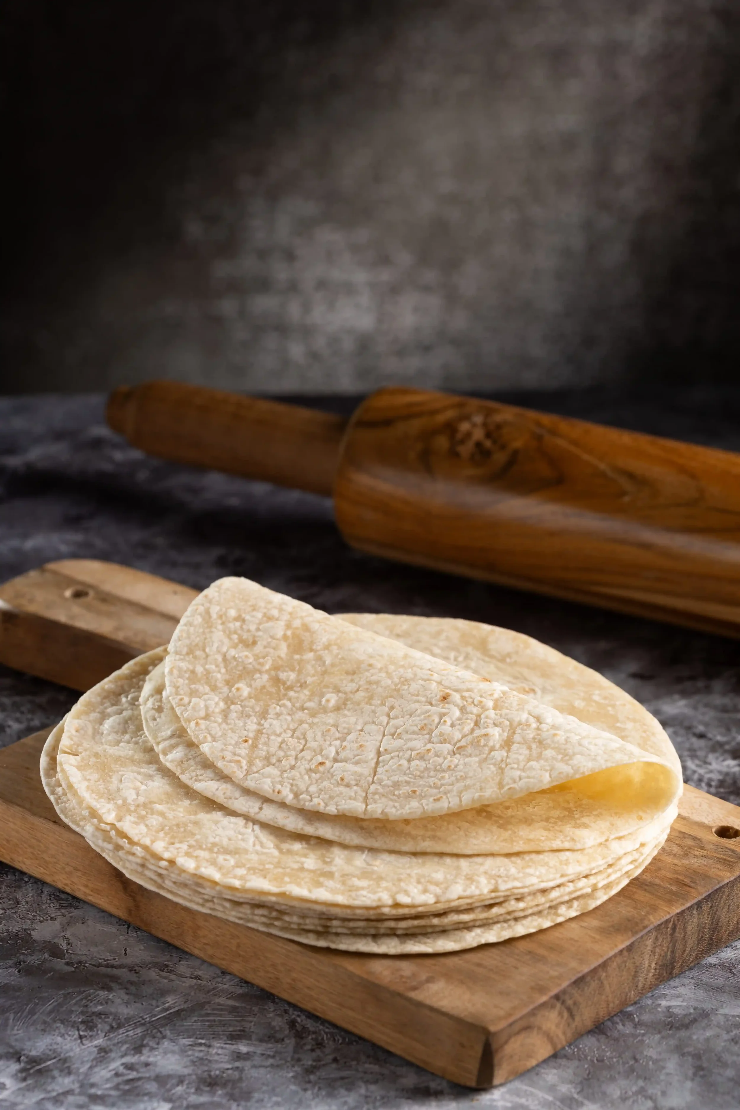 Stacked flour tortillas on a wooden cutting board, with a rolling pin in the background on a dark textured surface.