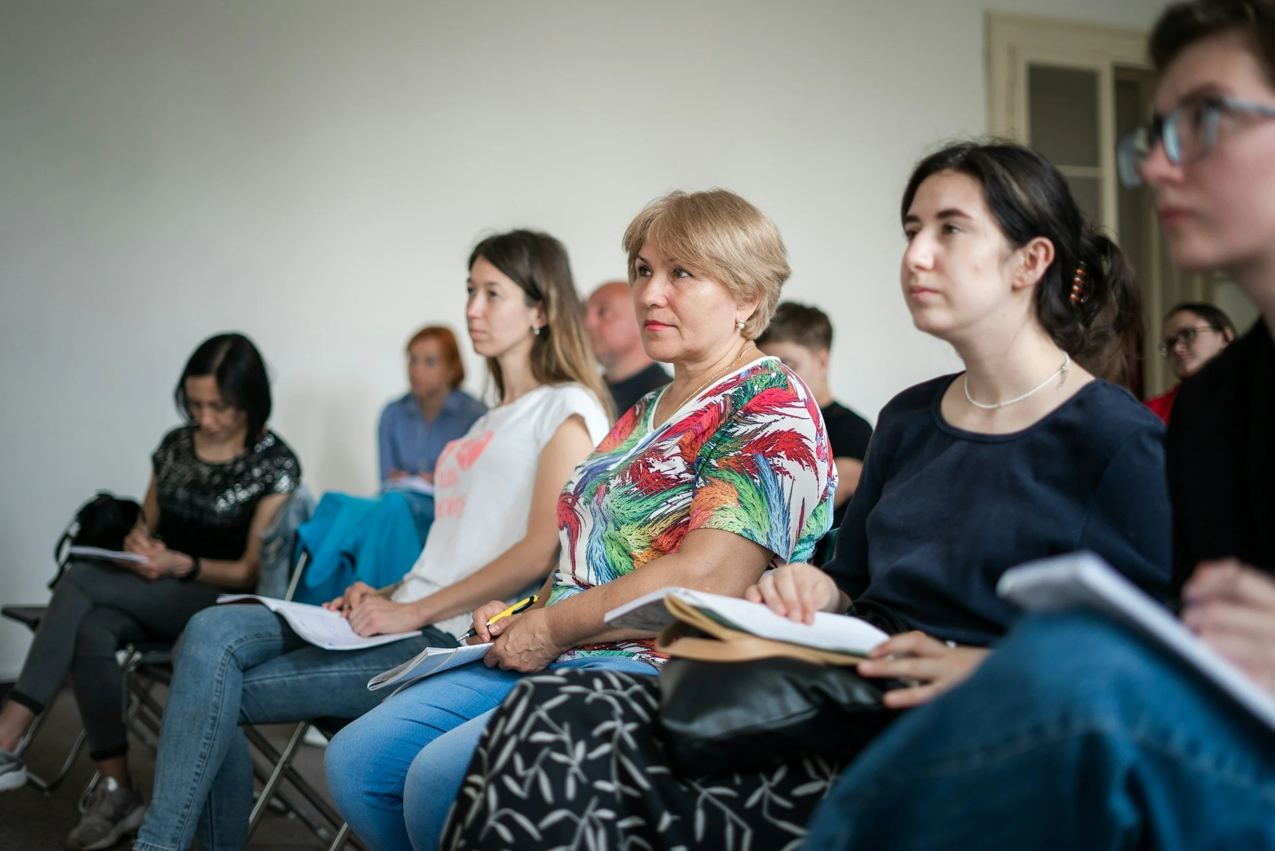 People seated in a meeting or conference, listening attentively, with notebooks and pens, in a well-lit room.