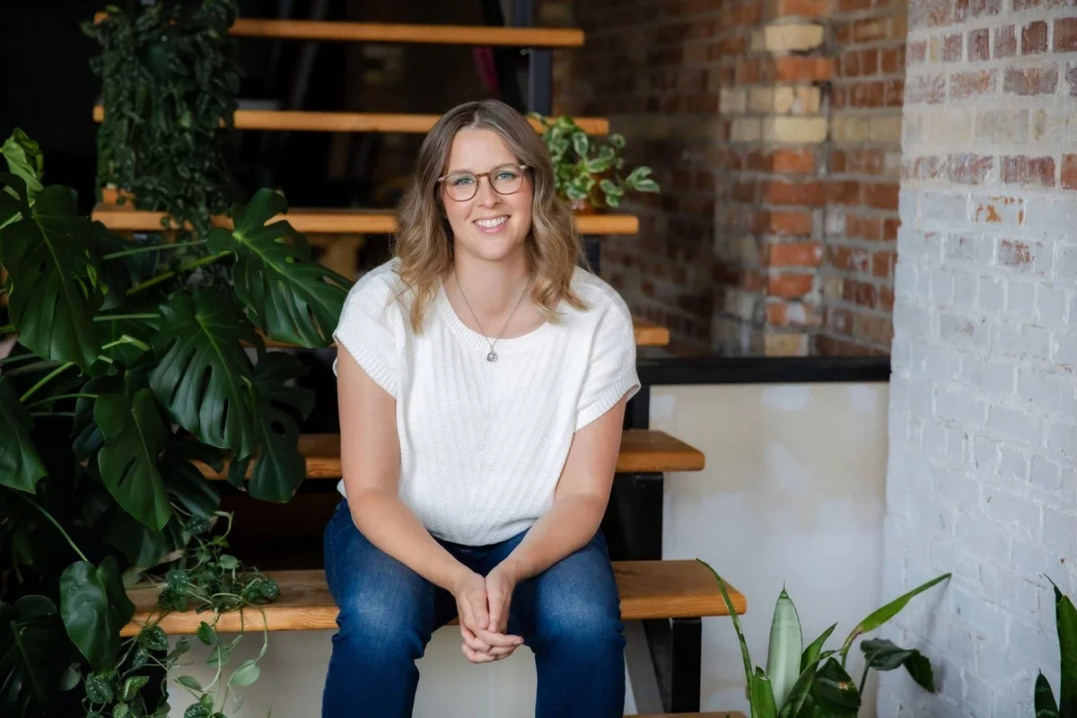 Megan Welwood smiling while sitting on wooden stairs beside green tropical plants and a brick wall.