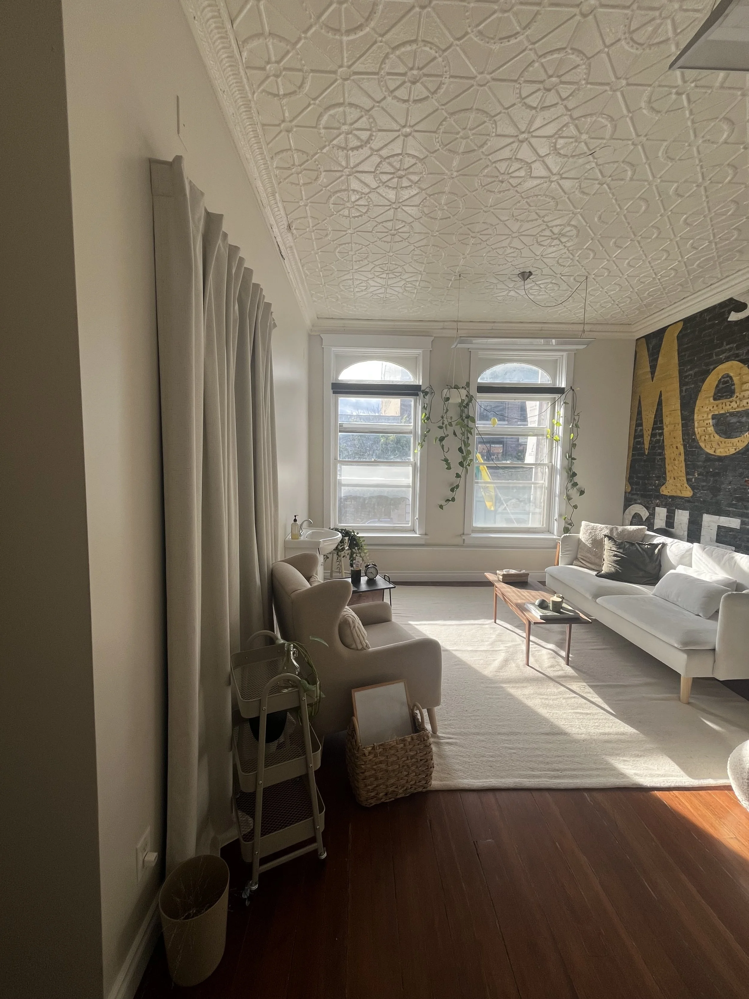Cozy counselling office space with white walls and ceiling, two large windows with white trim, a light gray couch with pillows, a small side table with various items, a hanging plant, and a wall with large decorative black and yellow letters.