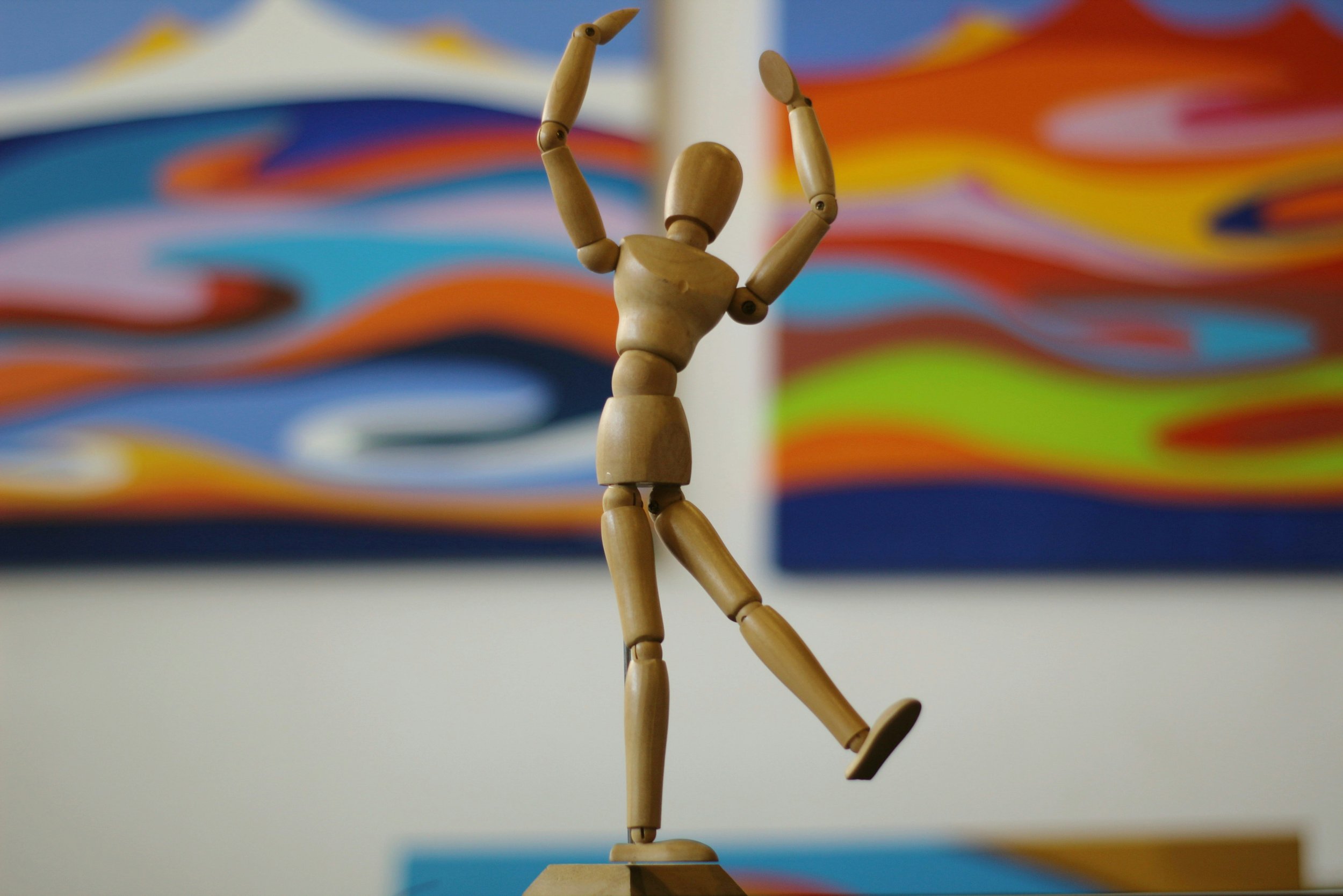 Wooden body drawing figure dummy raising it's hands in the air in front of a colourful wavy whimsical background.