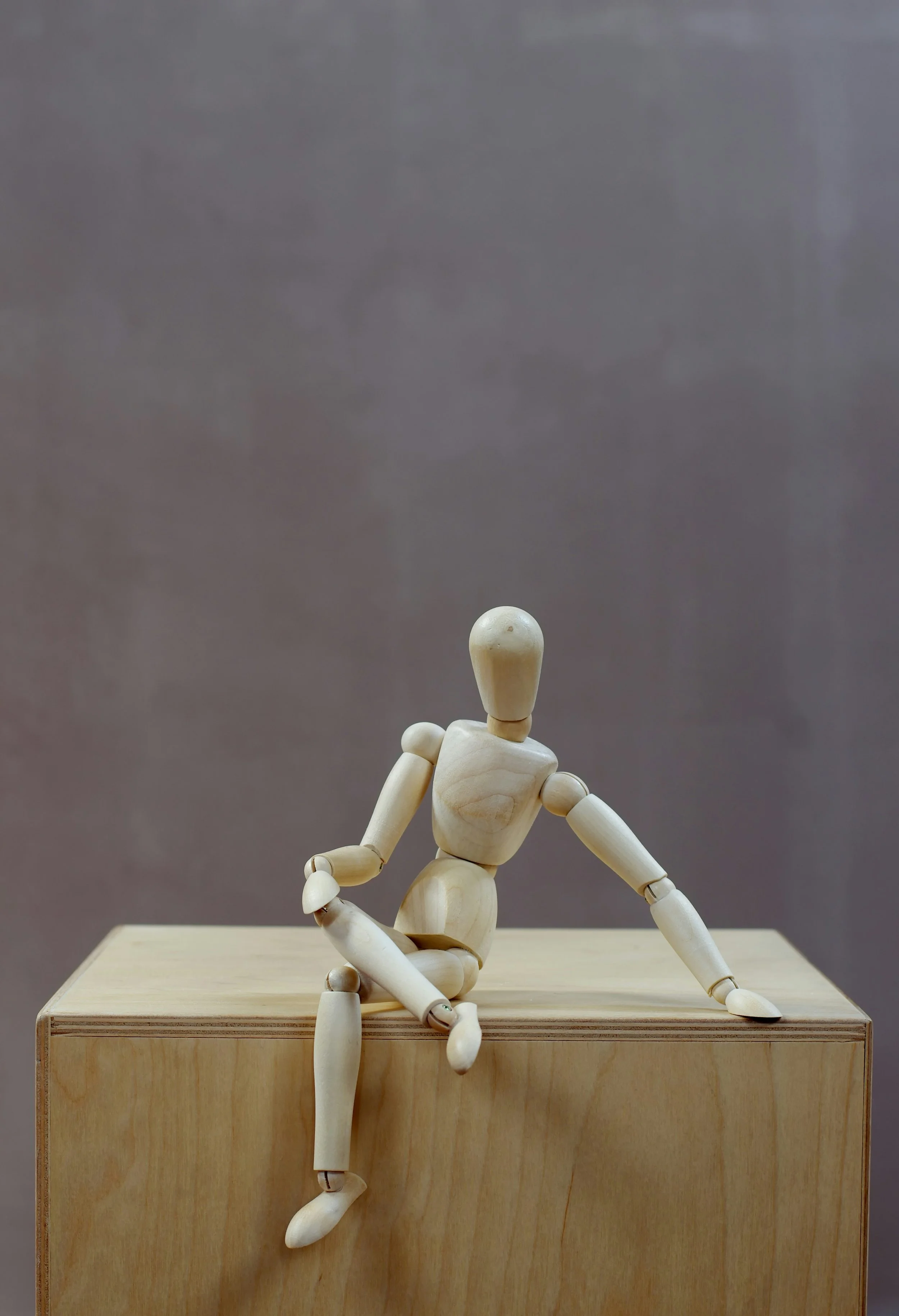 Wooden body dummy drawing figure sitting confidently on a wooden box in front of a grey background.