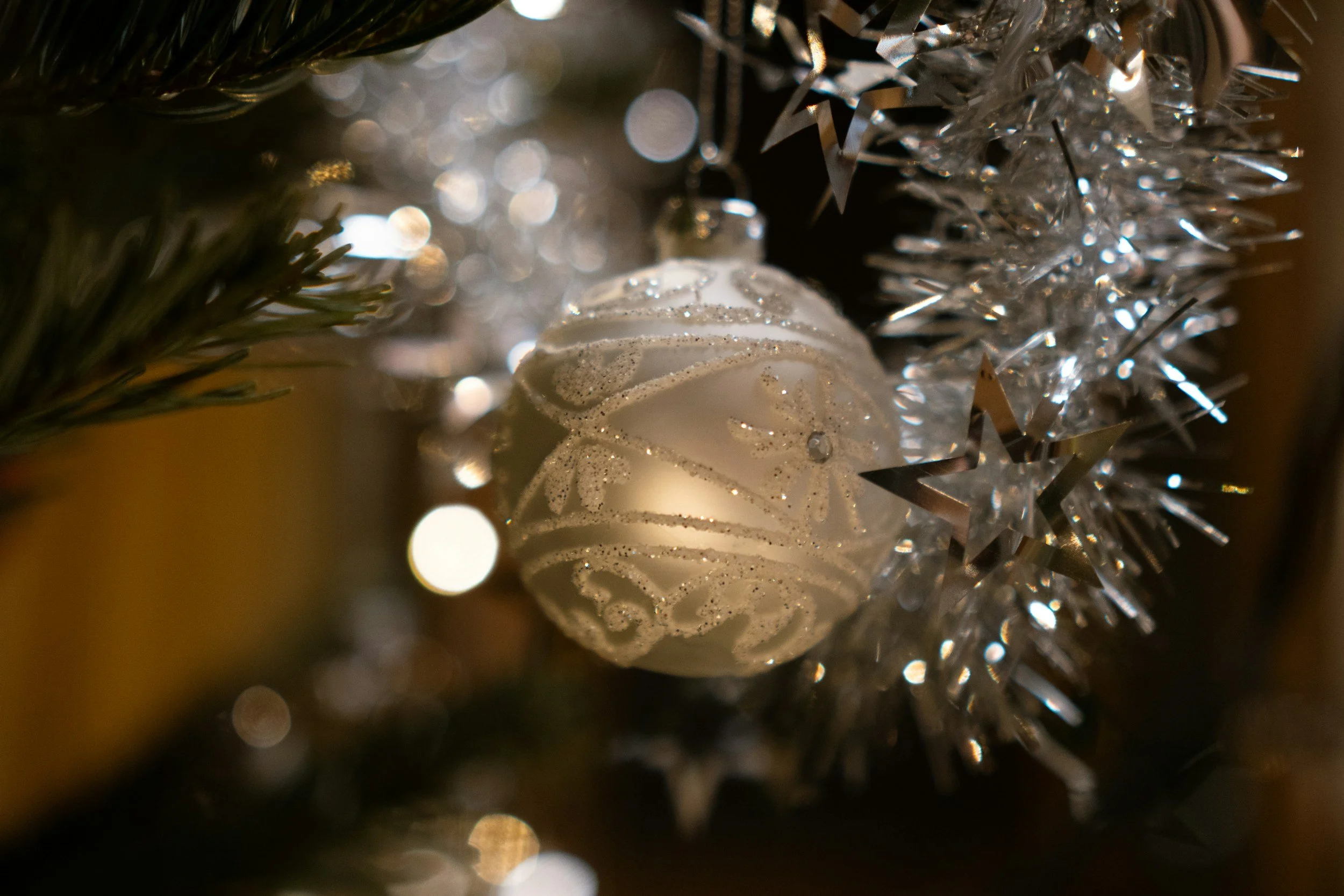Close-up of a white Christmas ornament with silver glitter and swirl designs, hanging on a Christmas tree decorated with silver tinsel and star-shaped ornaments.