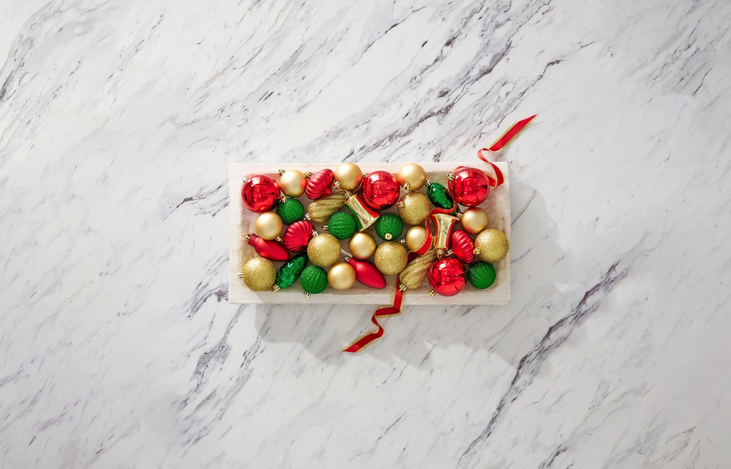 A white box filled with assorted Christmas ornaments in red, gold, and green, placed on a white marble surface with gray veining.