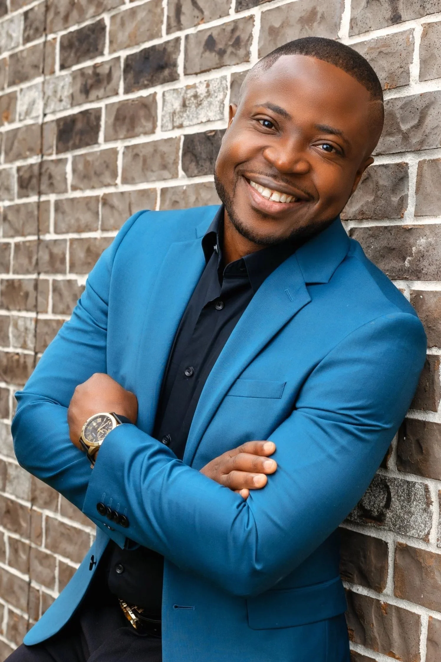 A smiling man wearing a blue blazer and black shirt with his arms crossed, leaning against a brick wall with grass at the bottom.