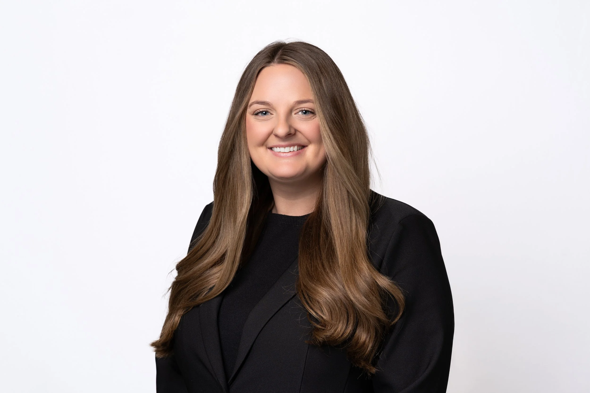 A woman with long light brown hair and fair skin, wearing a black blazer and black top, smiling against a plain white background.