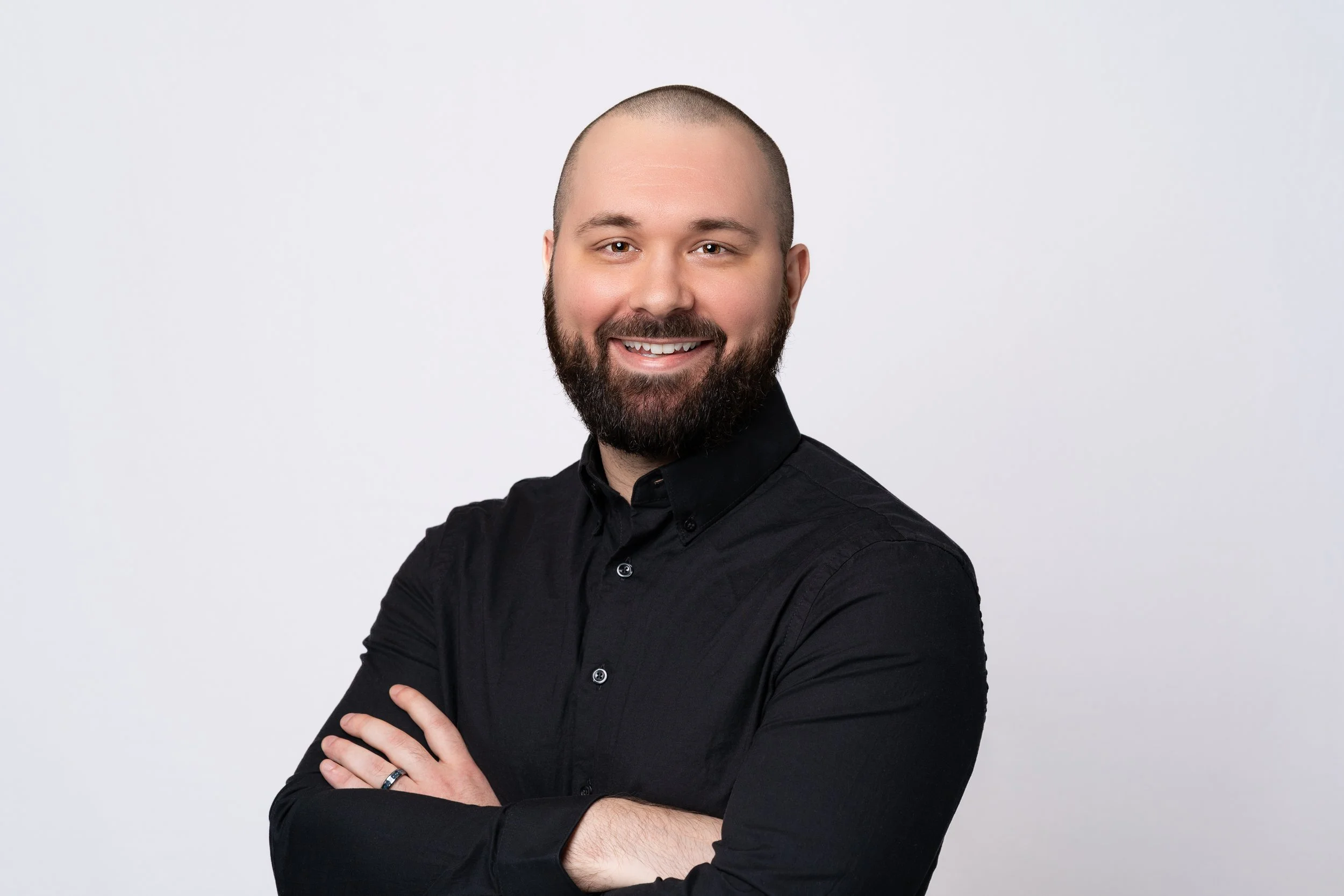 A man with a beard and short hair smiling with arms crossed, wearing a black shirt, against a plain light gray background.