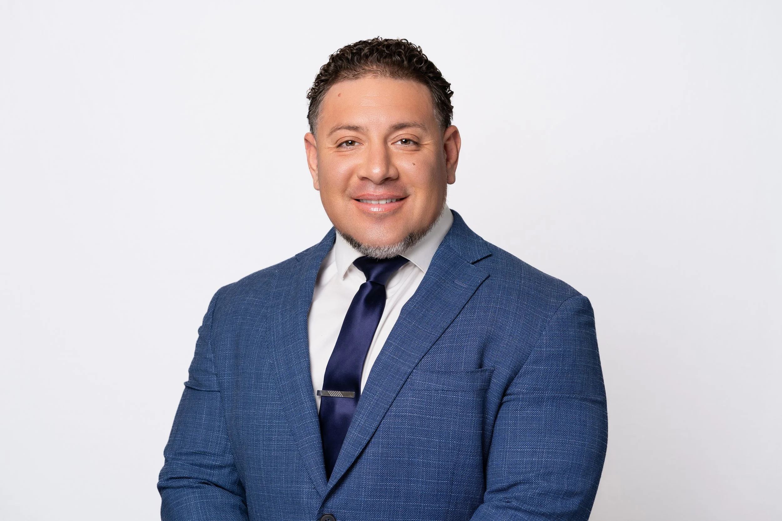 A man wearing a blue suit jacket, white shirt, and dark tie looking at the camera and smiling, with short, curly hair and a goatee, against a plain white background.