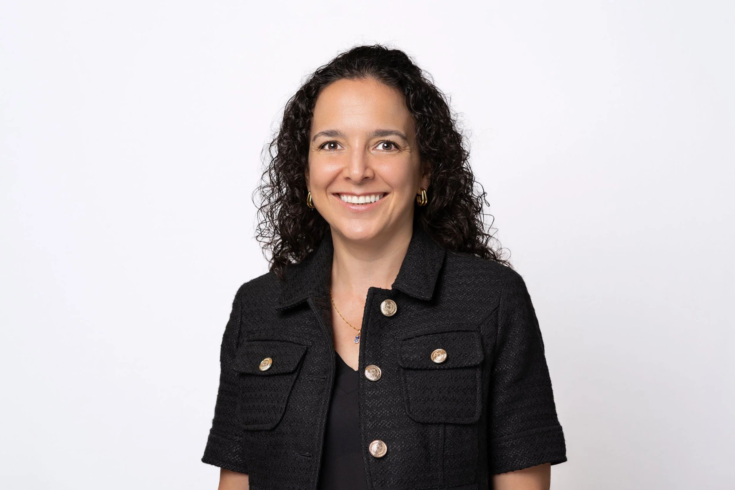 Portrait of a woman with curly dark hair wearing a black textured jacket with gold buttons and gold hoop earrings, smiling against a plain white background.