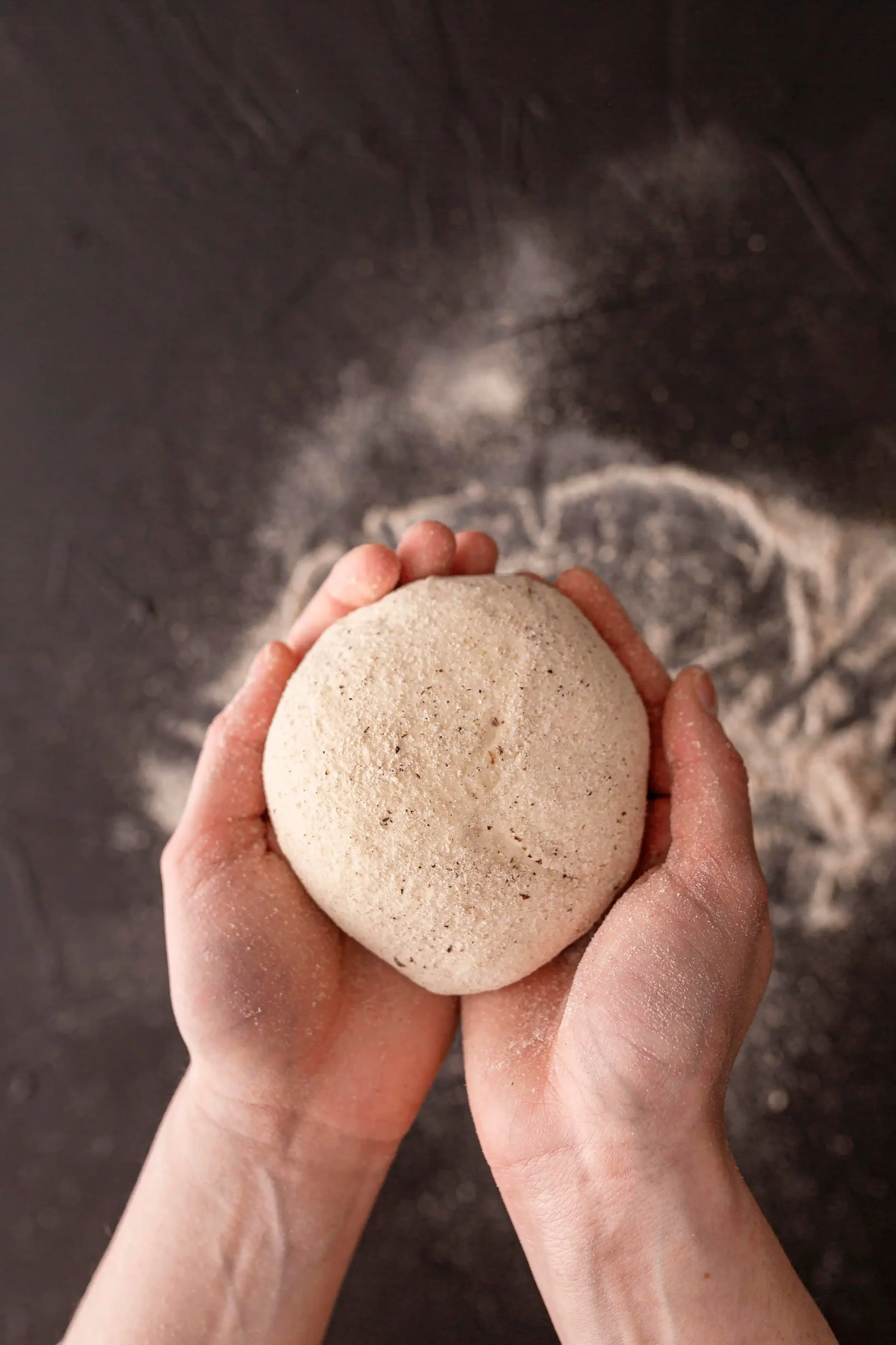 A person holding a round ball of dough with both hands above a baking sheet with flour dusted on it.