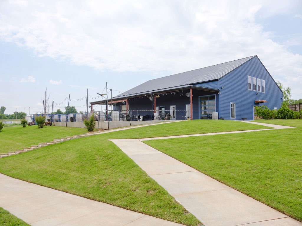 A blue barn-style building with a sloped metal roof sits on a grassy hill. A concrete path leads up to the building, which features an outdoor patio area with string lights and seating. The sky is partly cloudy.