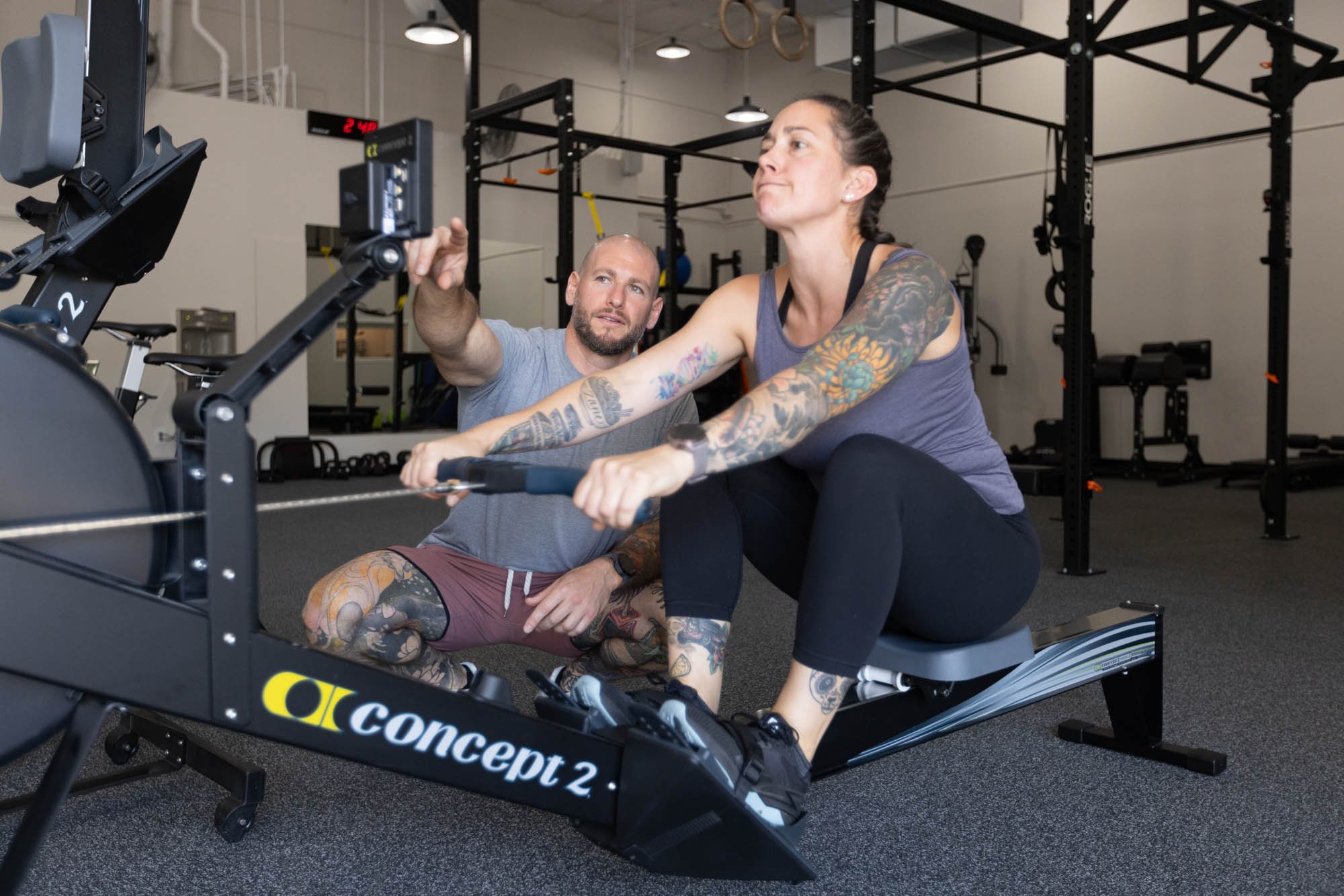 Woman exercising on rowing machine with personal trainer's guidance.