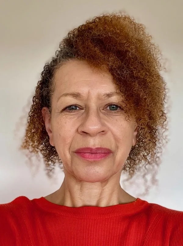 Maggy Burrowes. Portrait of a woman with curly hair, wearing a red top, against a plain background.