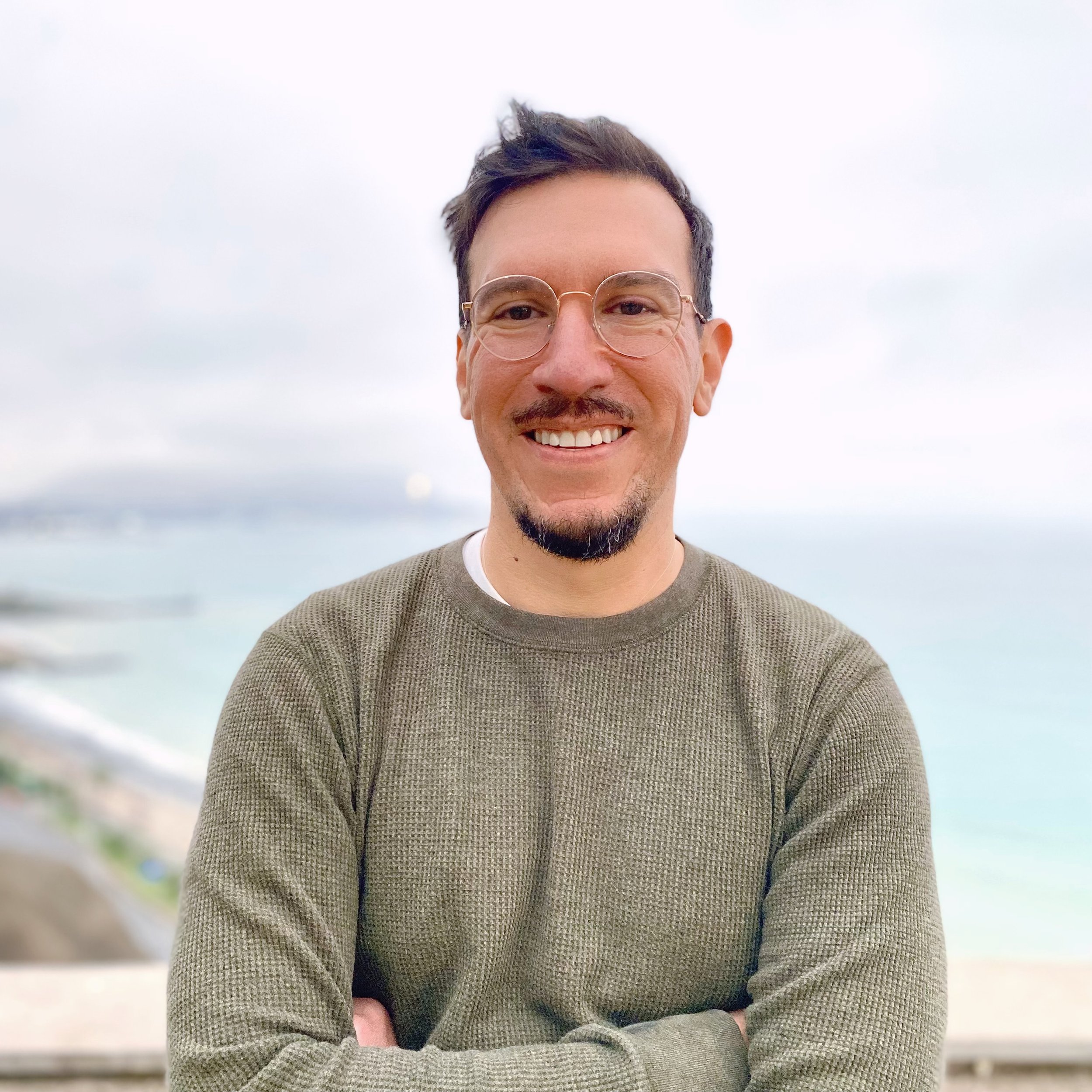Smiling man with glasses and a beard, wearing a green sweater, standing outdoors by the water.