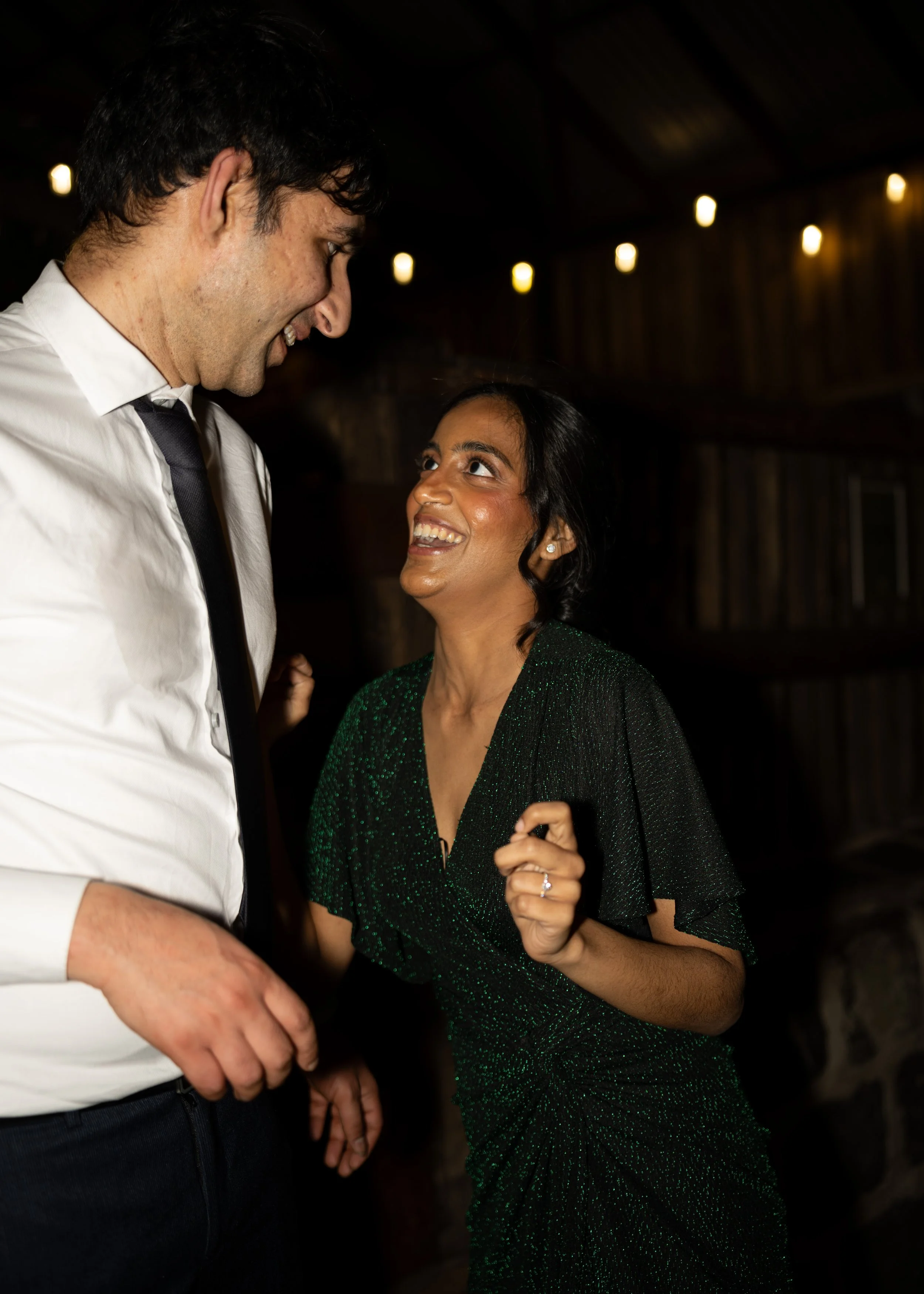 A man and woman are smiling and looking at each other at a social event, with warm lighting and wooden interior background.