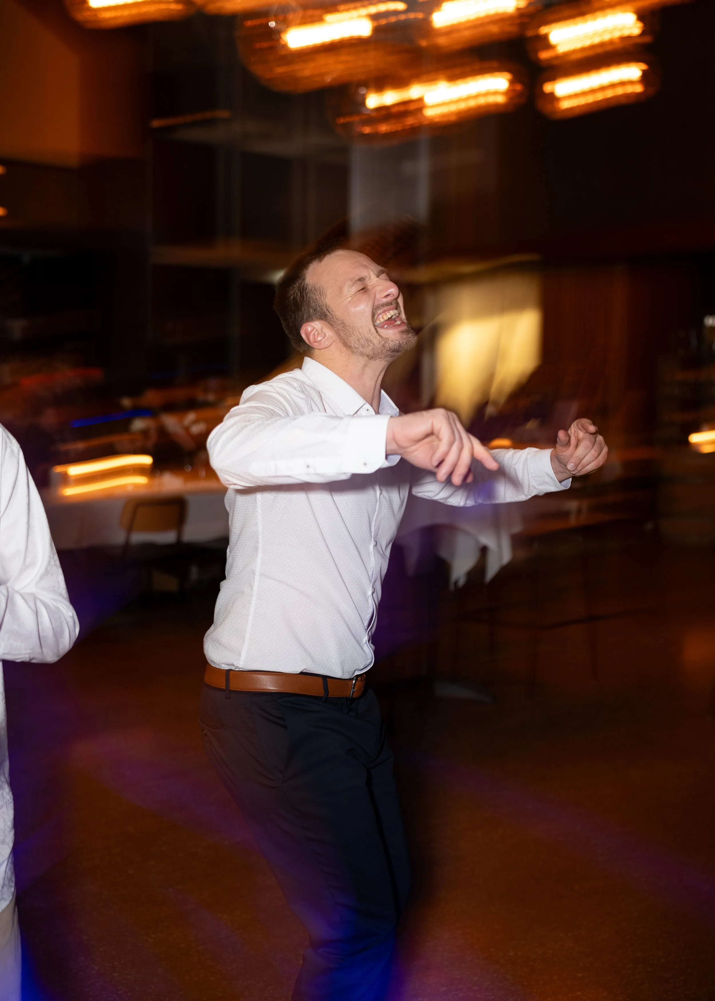 A man dancing joyfully at a party or celebration, wearing a white shirt and dark pants, with blurred background lights.