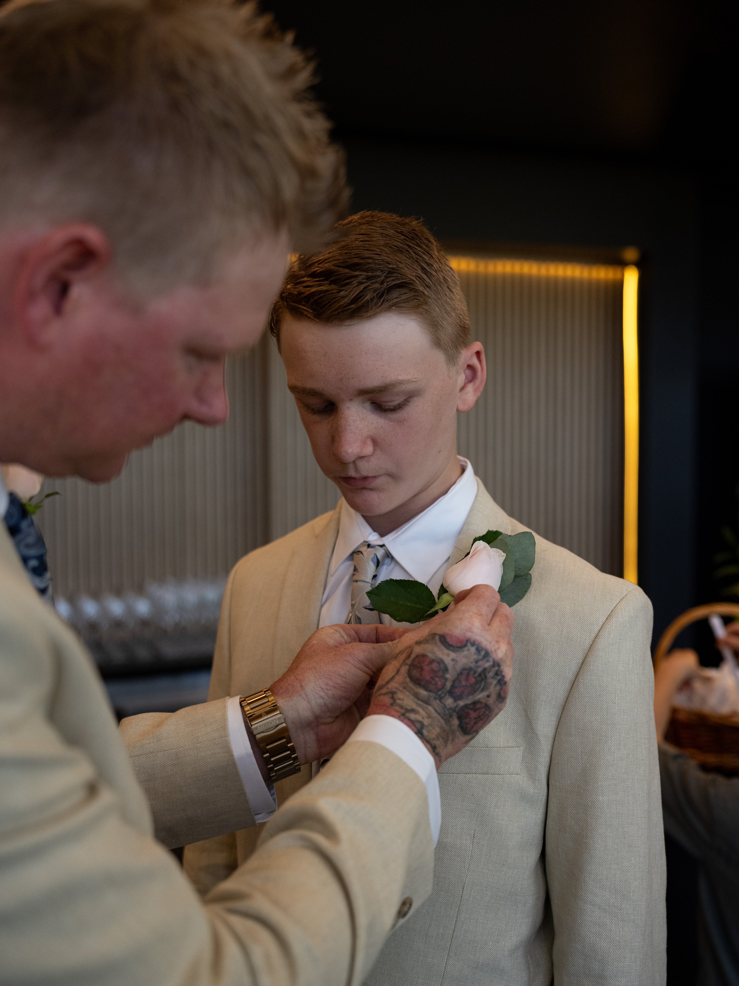 A man pins a white rose boutonniere on a young man's lapel during a formal event.