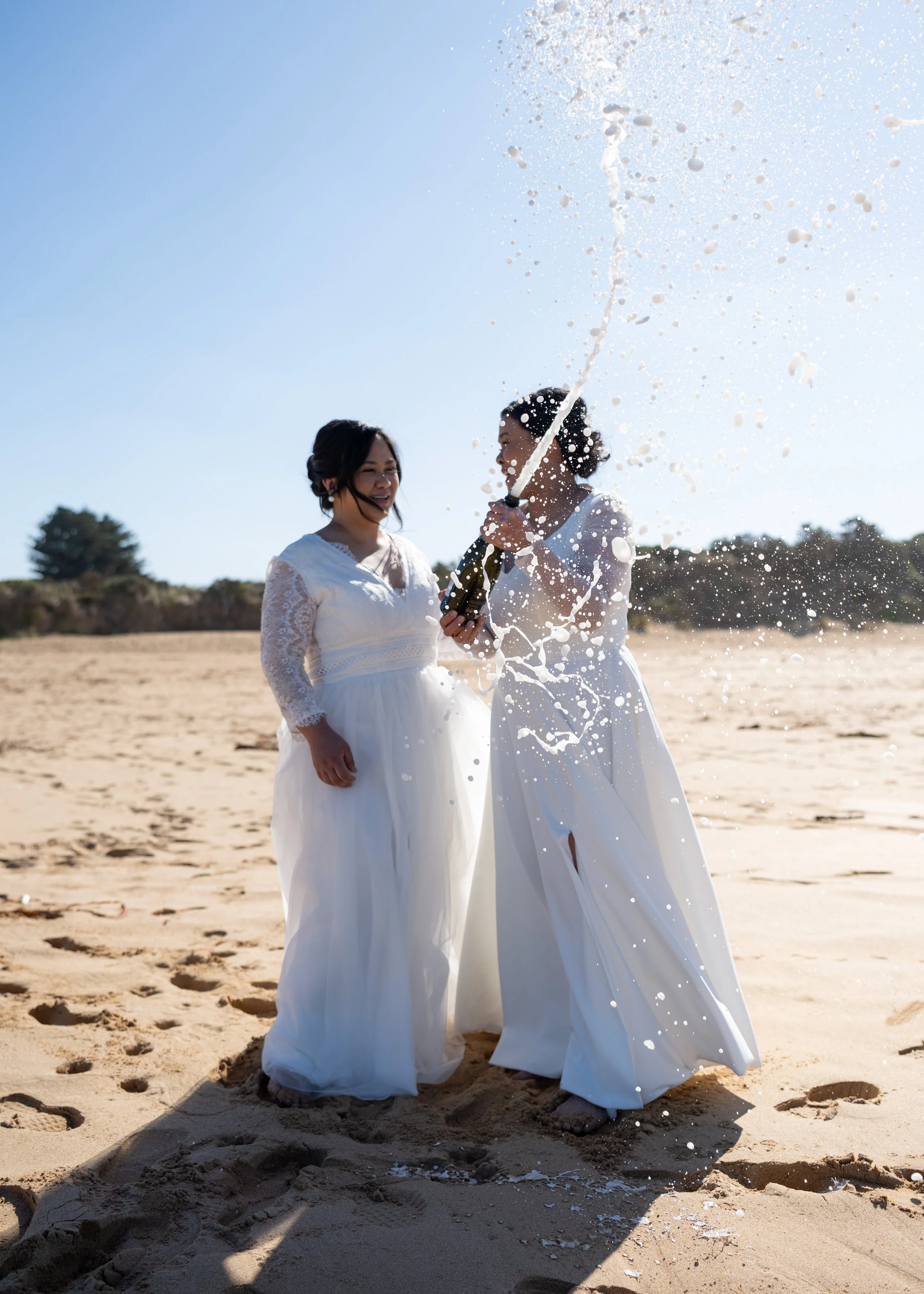 Two women in white dresses celebrating with champagne on a sandy beach, one opening a bottle and spraying it in the air, under a clear blue sky.