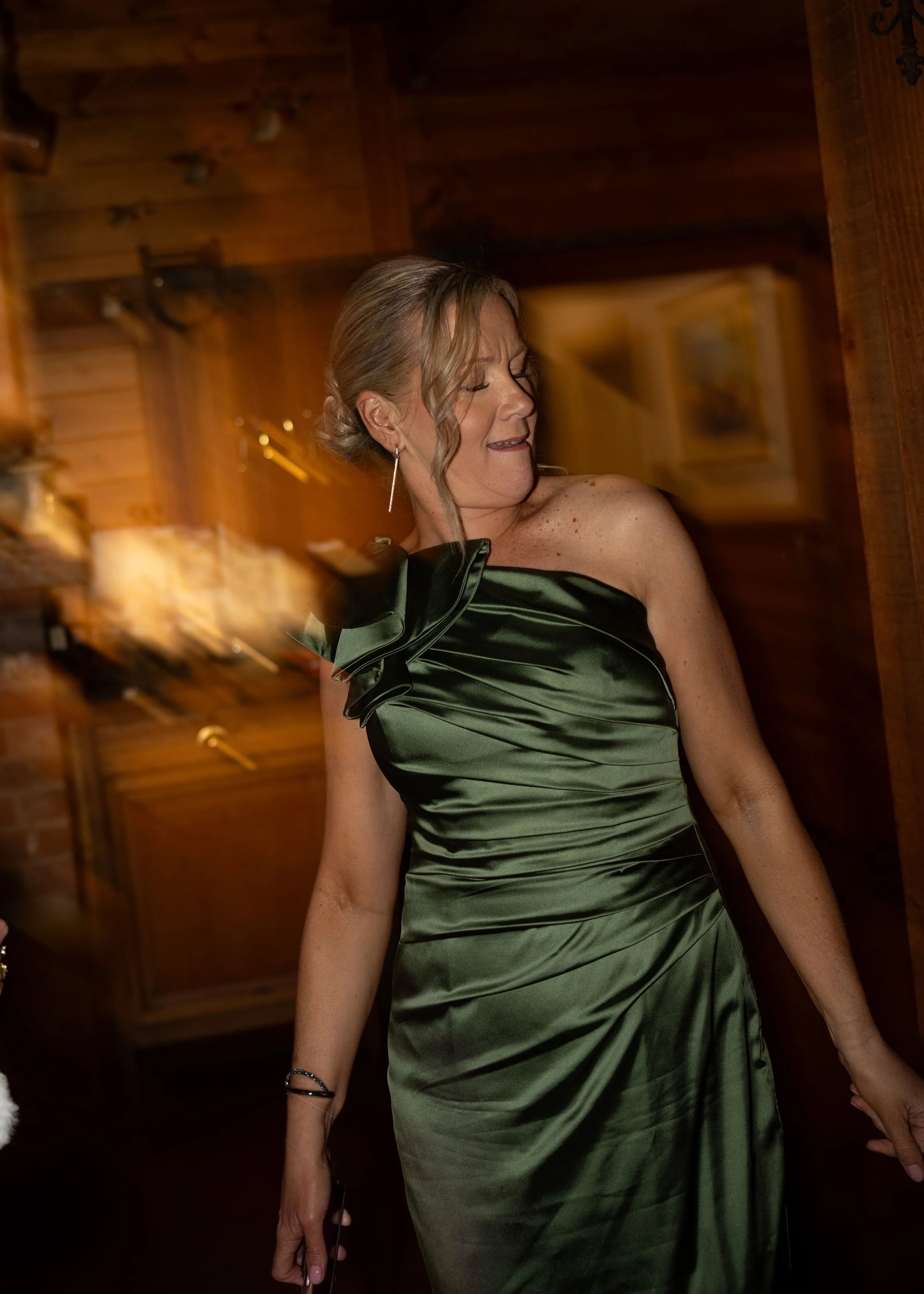A woman in an elegant dark green satin dress with a large bow detail on the shoulder, standing in a wooden room, with short curly hair and earrings, appearing to enjoy herself.