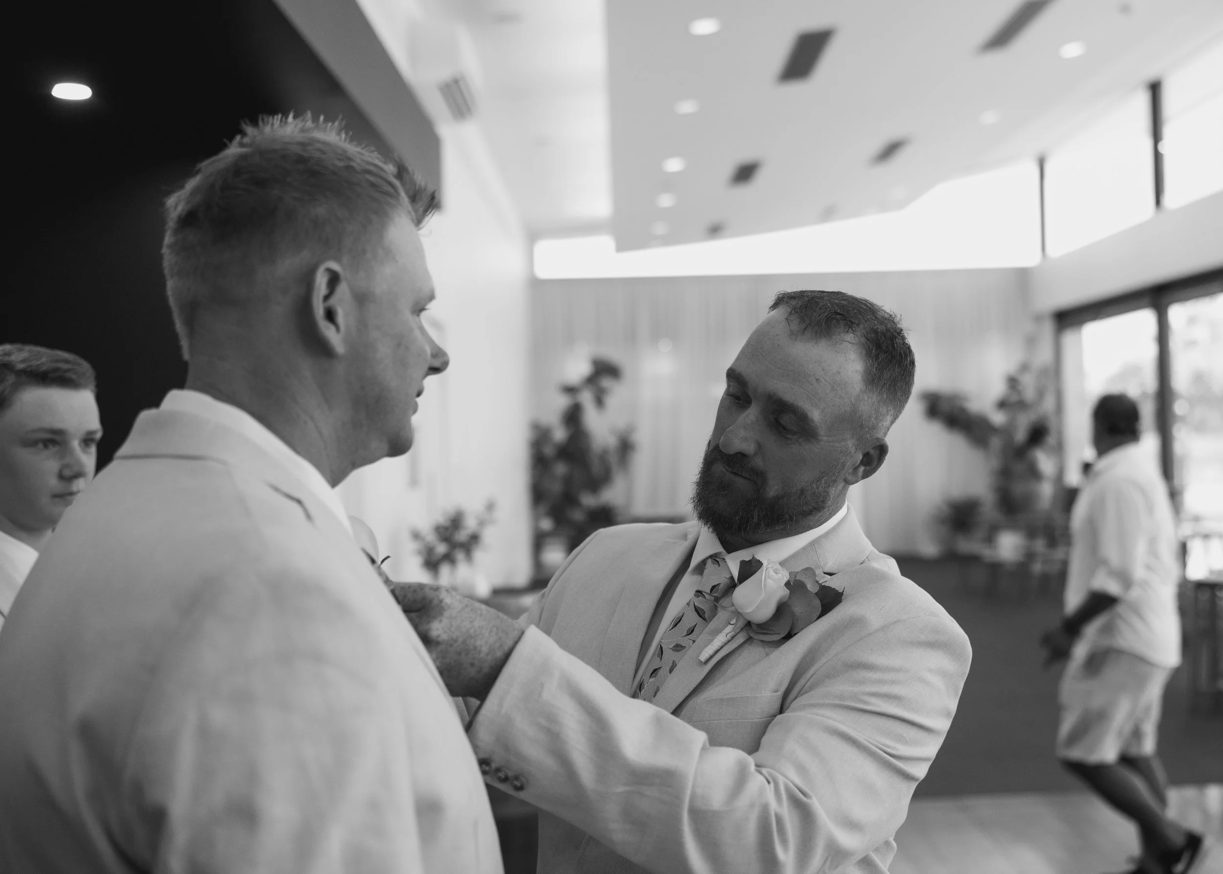 Black-and-white photo of two men in suits, one adjusting the other's tie, in a bright indoor setting. A boy and a woman are in the background, with large windows and plants visible.