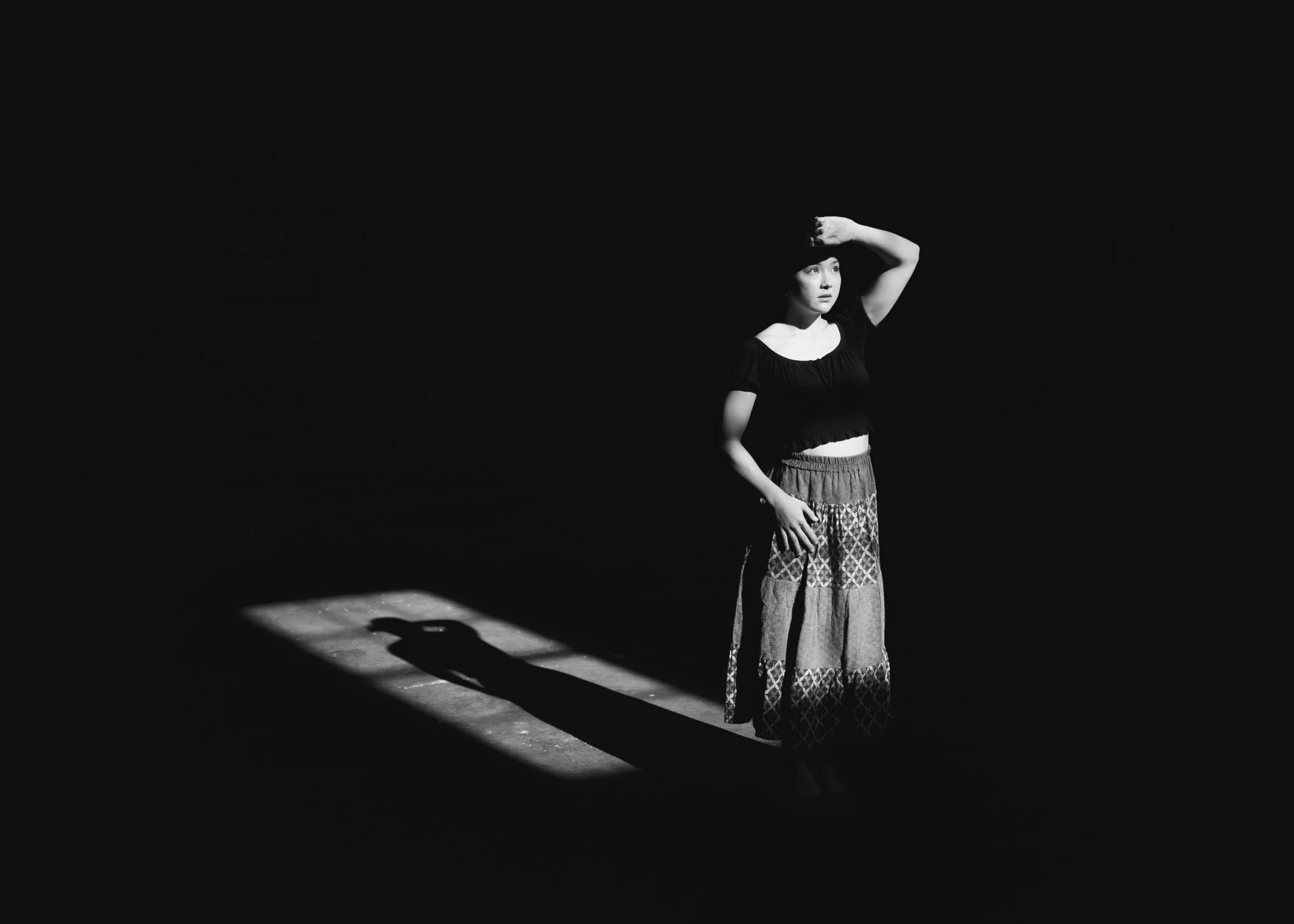 A woman standing alone in a dark space, illuminated by a single spotlight. She has her right hand on her forehead and wears a black top with short sleeves and a long, patterned skirt. Her shadow is cast on the floor.