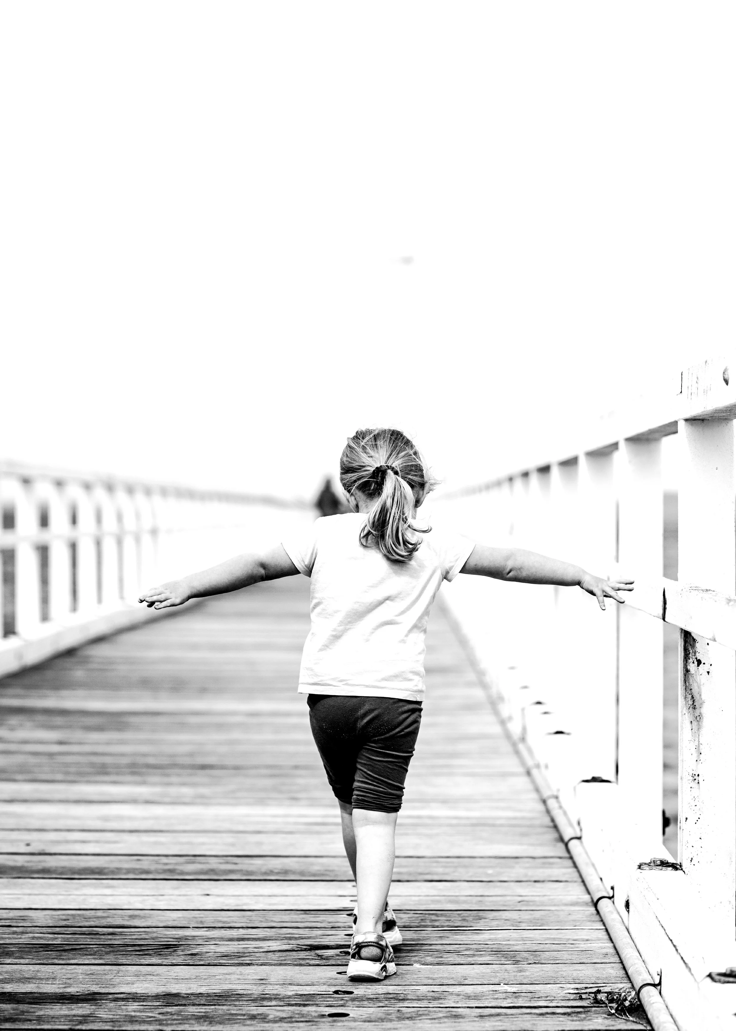A black-and-white photo of a young girl walking on a wooden bridge with her arms outstretched.