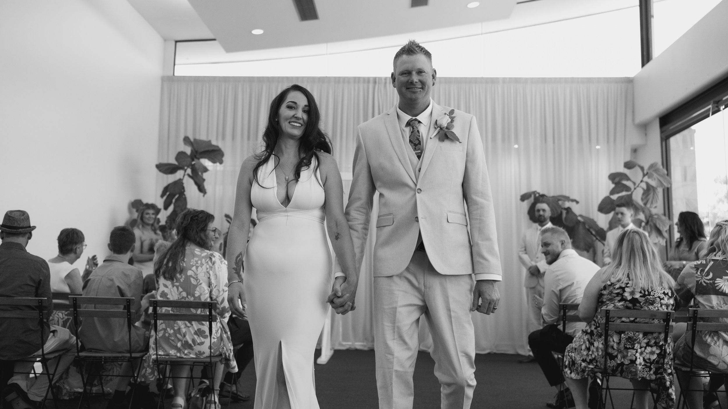 A couple at their wedding, holding hands and smiling, walking down the aisle with guests seated on either side in a decorated indoor venue.