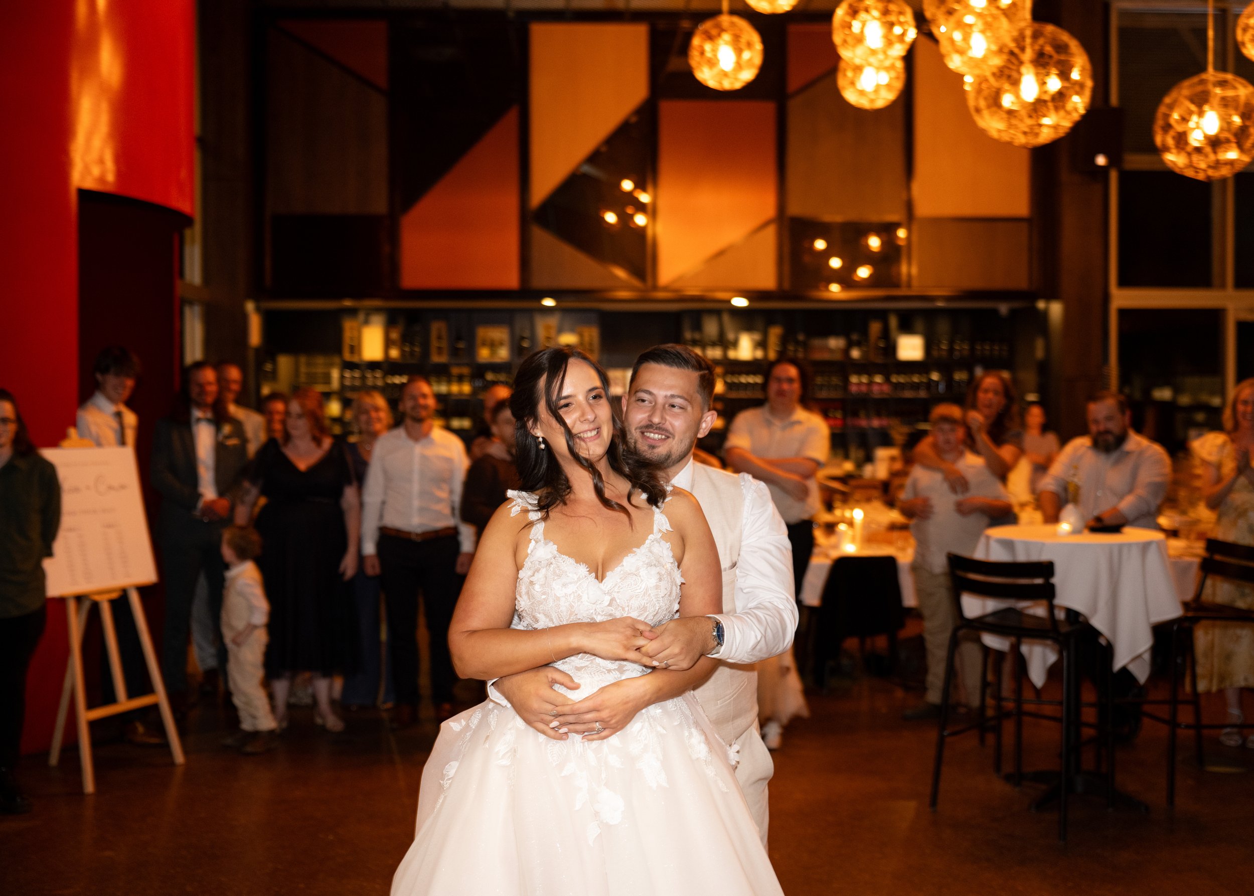 Bride and groom dancing at their wedding reception, surrounded by guests in an elegant, warmly lit venue.