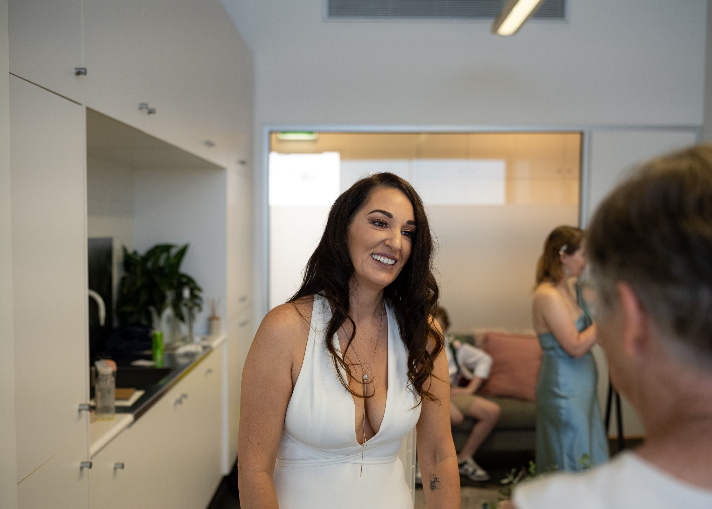 A woman smiling and talking to another person in an indoor setting, with several other people in the background.