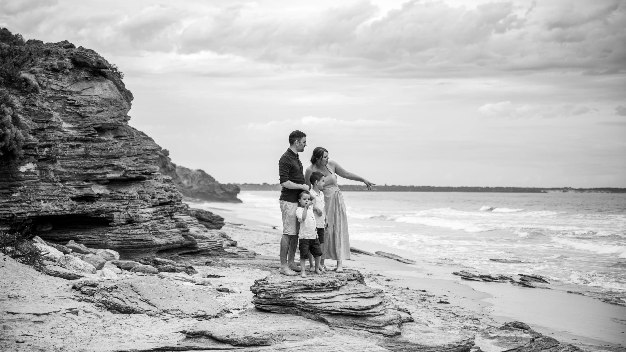 A family of four stands on a rock formation at the beach, with the ocean and cloudy sky in the background.