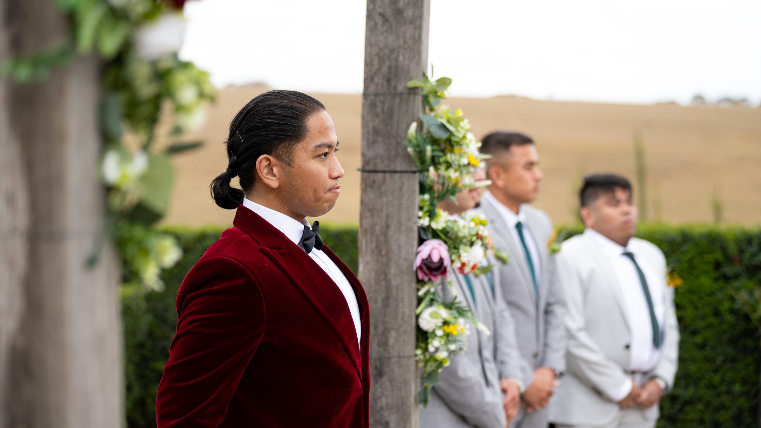 Man in a red velvet tuxedo standing next to a decorated wooden post during a ceremony, with three men in suits blurred in the background.
