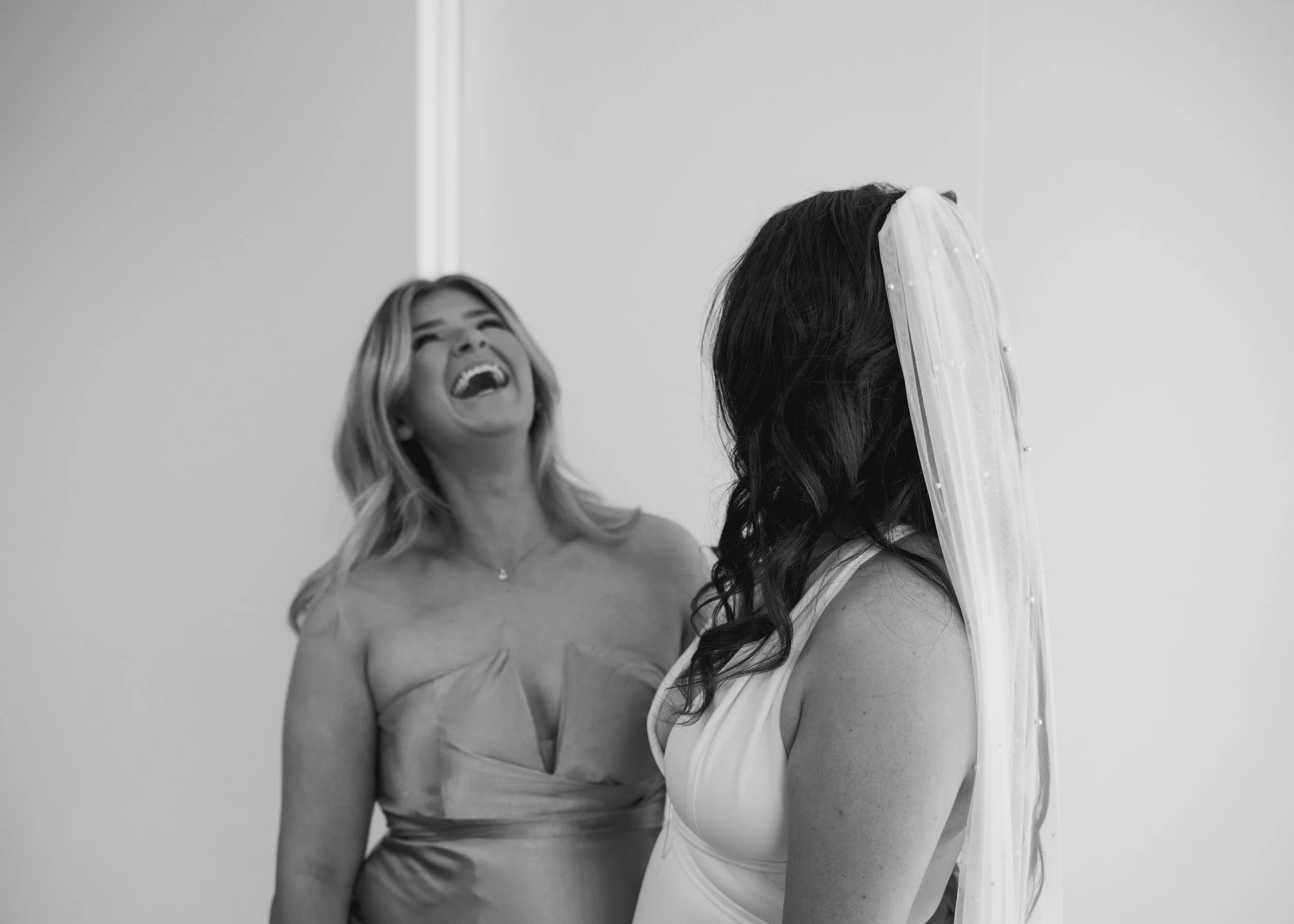 A woman in a wedding dress with a veil laughing with another woman in a sleeveless dress.