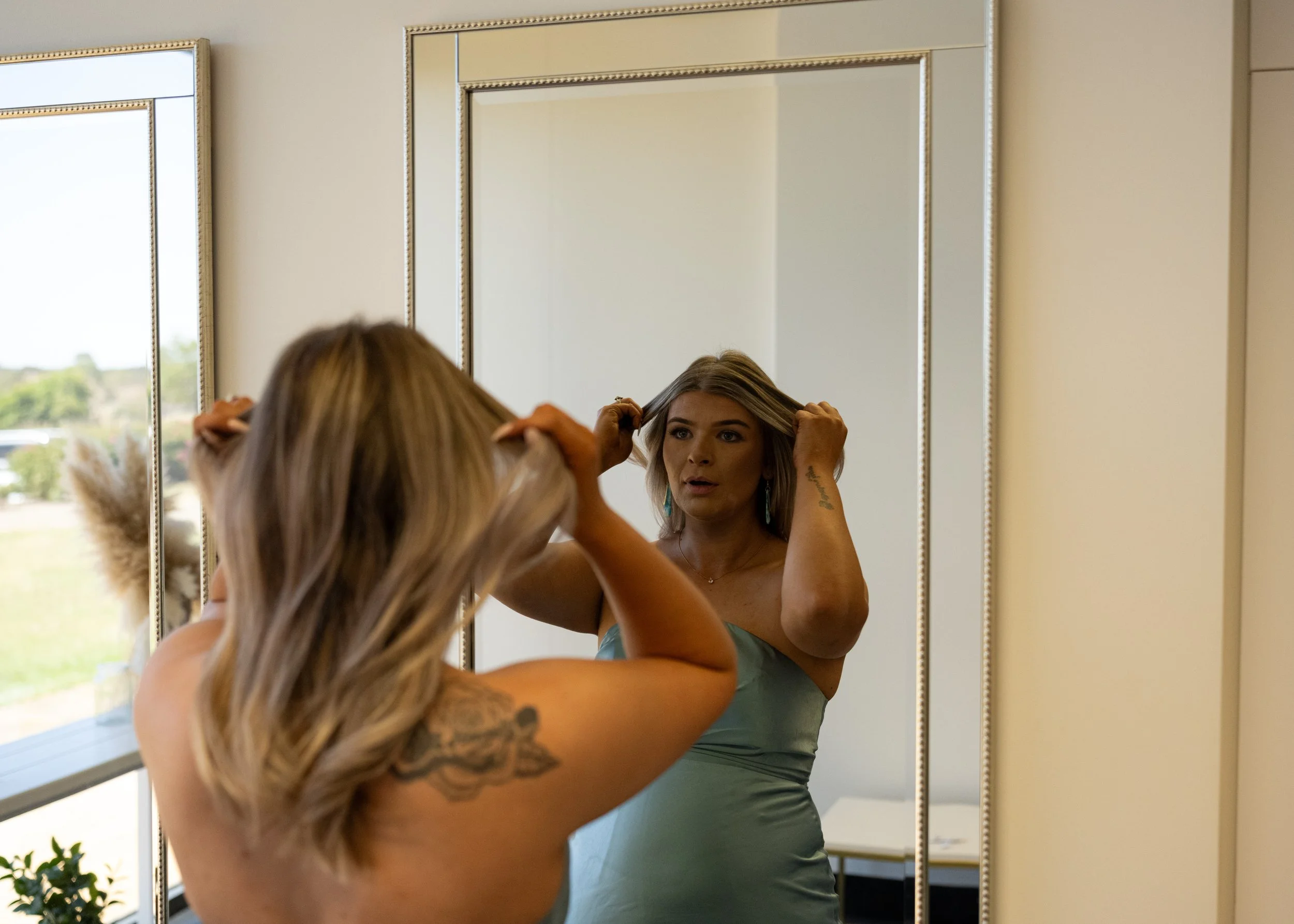A woman with blonde hair is adjusting her hair while looking in a mirror. She is wearing a strapless light blue dress and earrings. The mirror is positioned on a beige wall near a window with a view of outdoors.