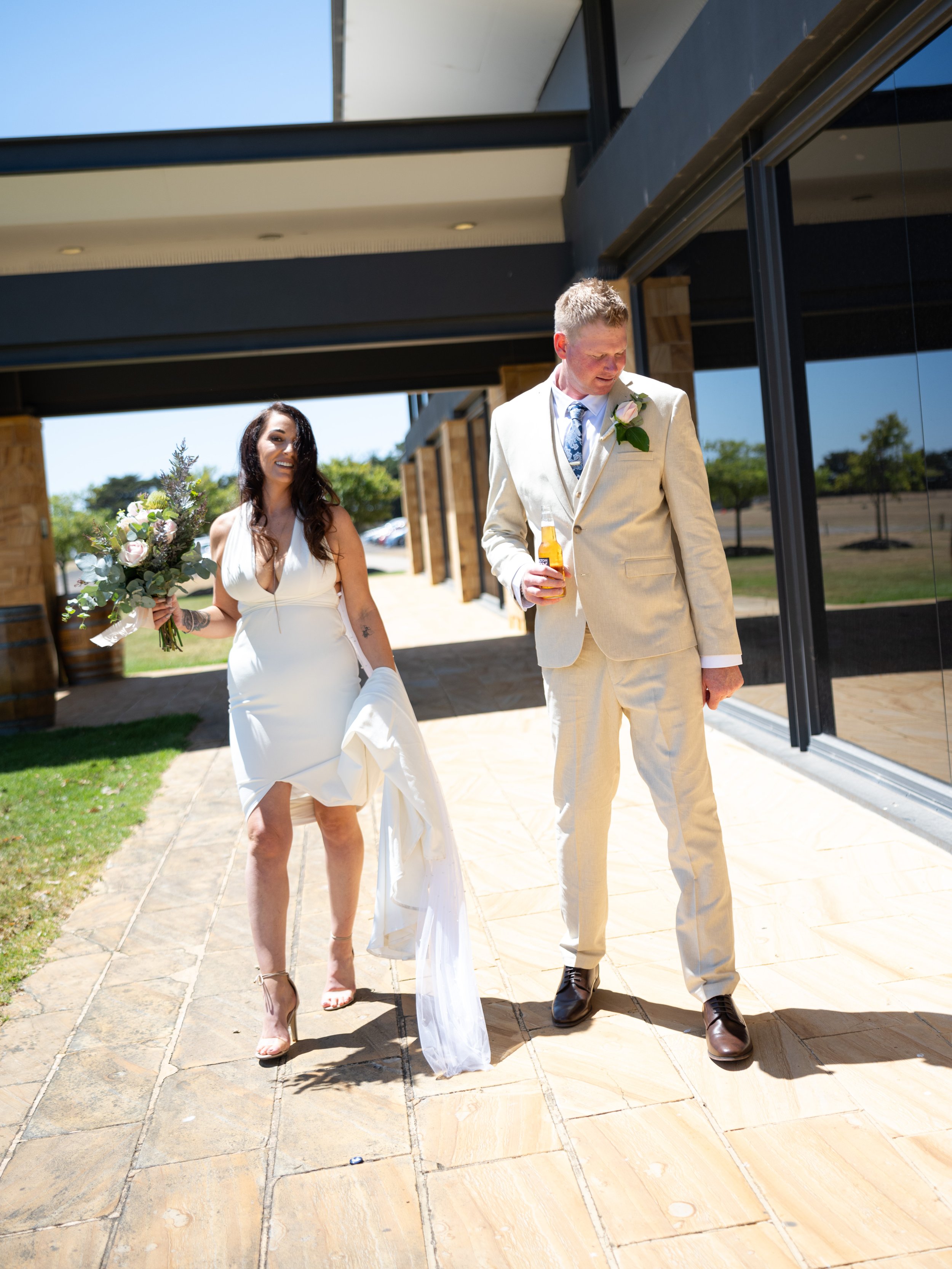 A bride and groom walking outside a modern building on a sunny day, with the bride holding a bouquet of flowers and the groom holding a bottle of beer.