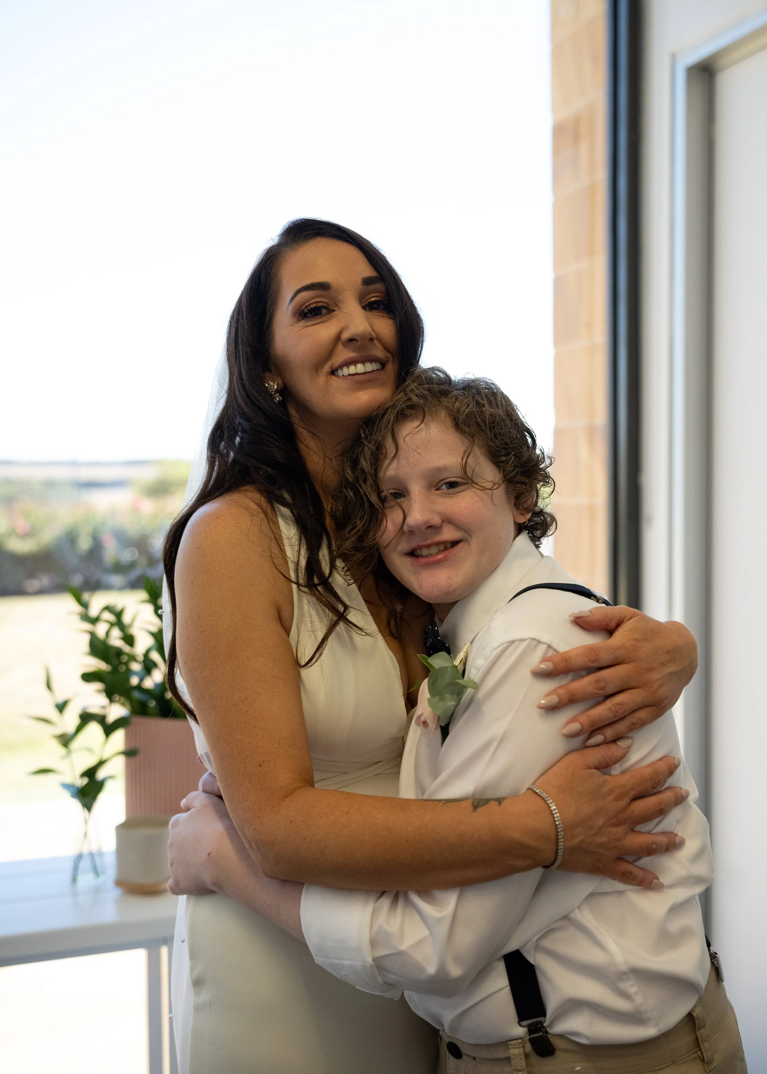 A woman in a wedding dress hugging a young boy in a white shirt and bow tie, both smiling, indoors near large windows with a view of greenery outside.