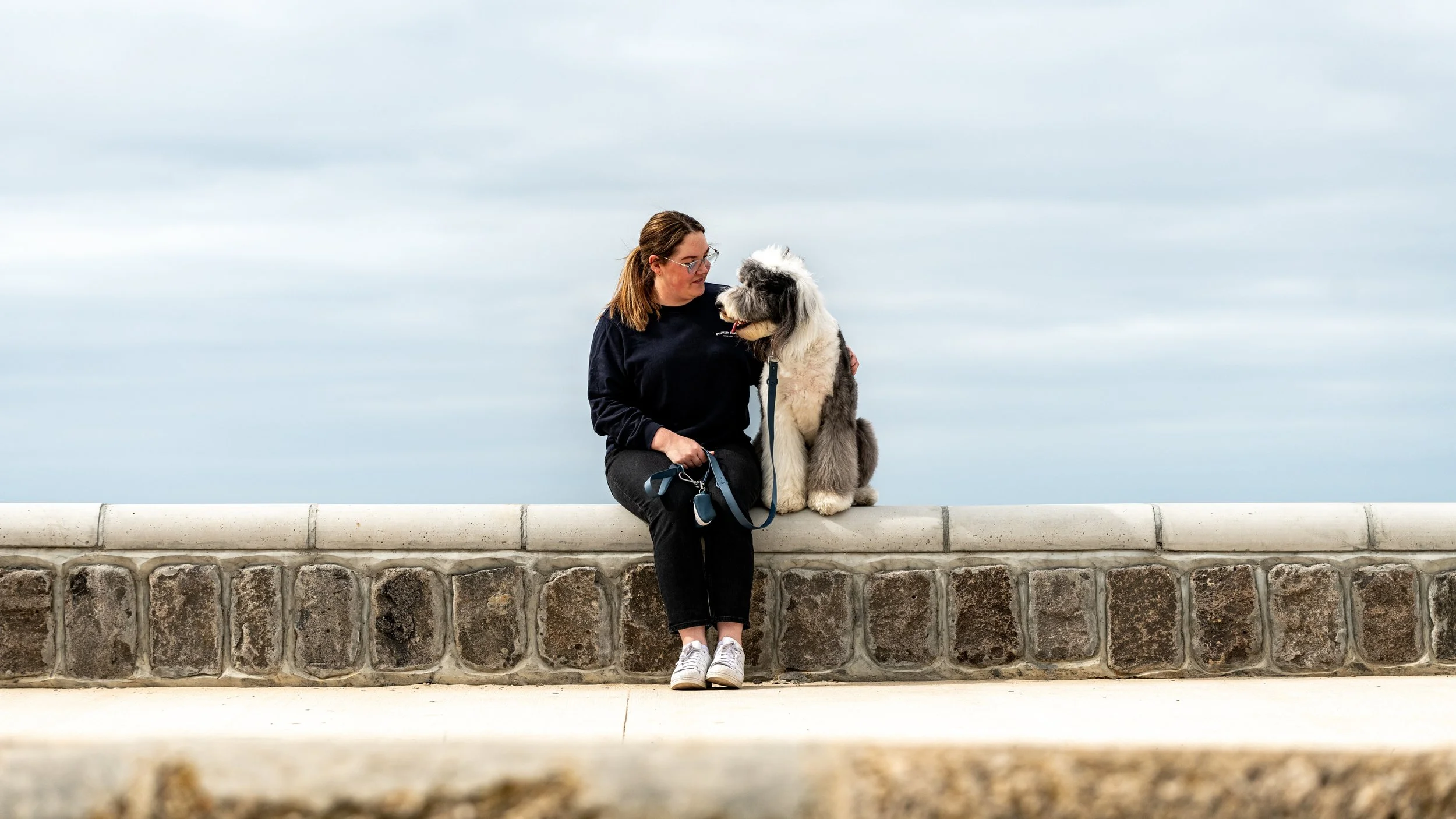 A woman sitting on a stone ledge with a large, fluffy dog, both looking at each other, under a cloudy sky.