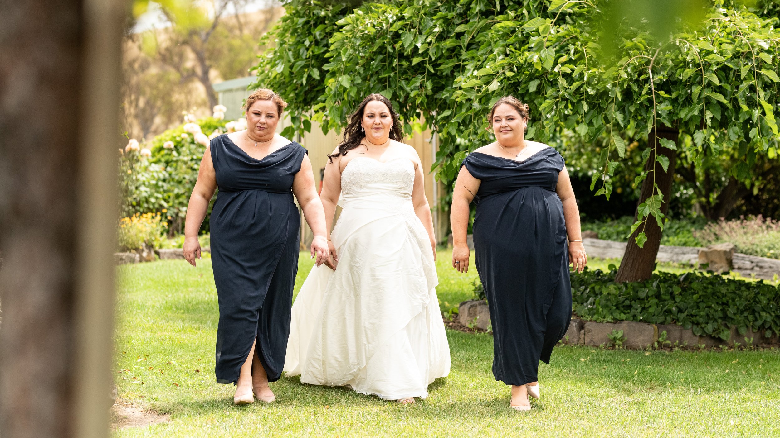 A bride walking with two bridesmaids under green trees on a grassy lawn.