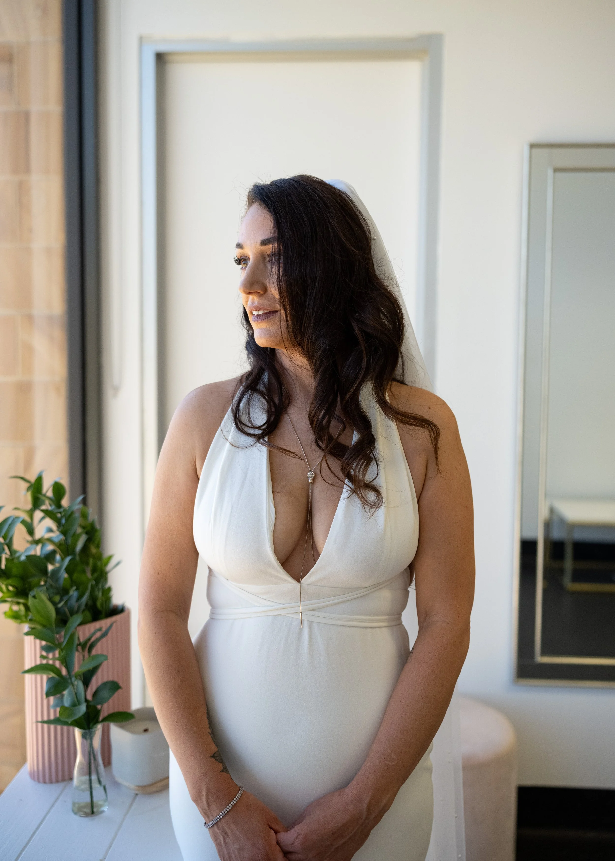 A woman in a white dress with dark brown hair stands indoors, looking out of a window with a soft smile, next to a pink vase with green plants.