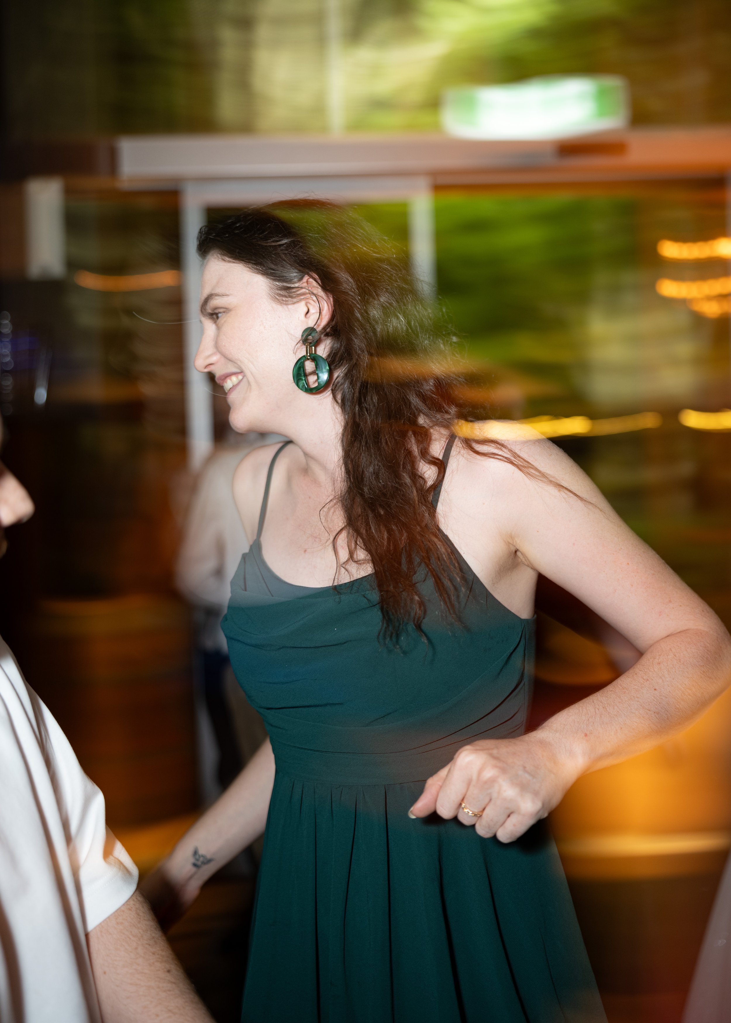 A woman with long dark hair wearing a dark green dress and large green earrings is smiling and engaged in conversation in a lively setting, with blurred colorful lights in the background.