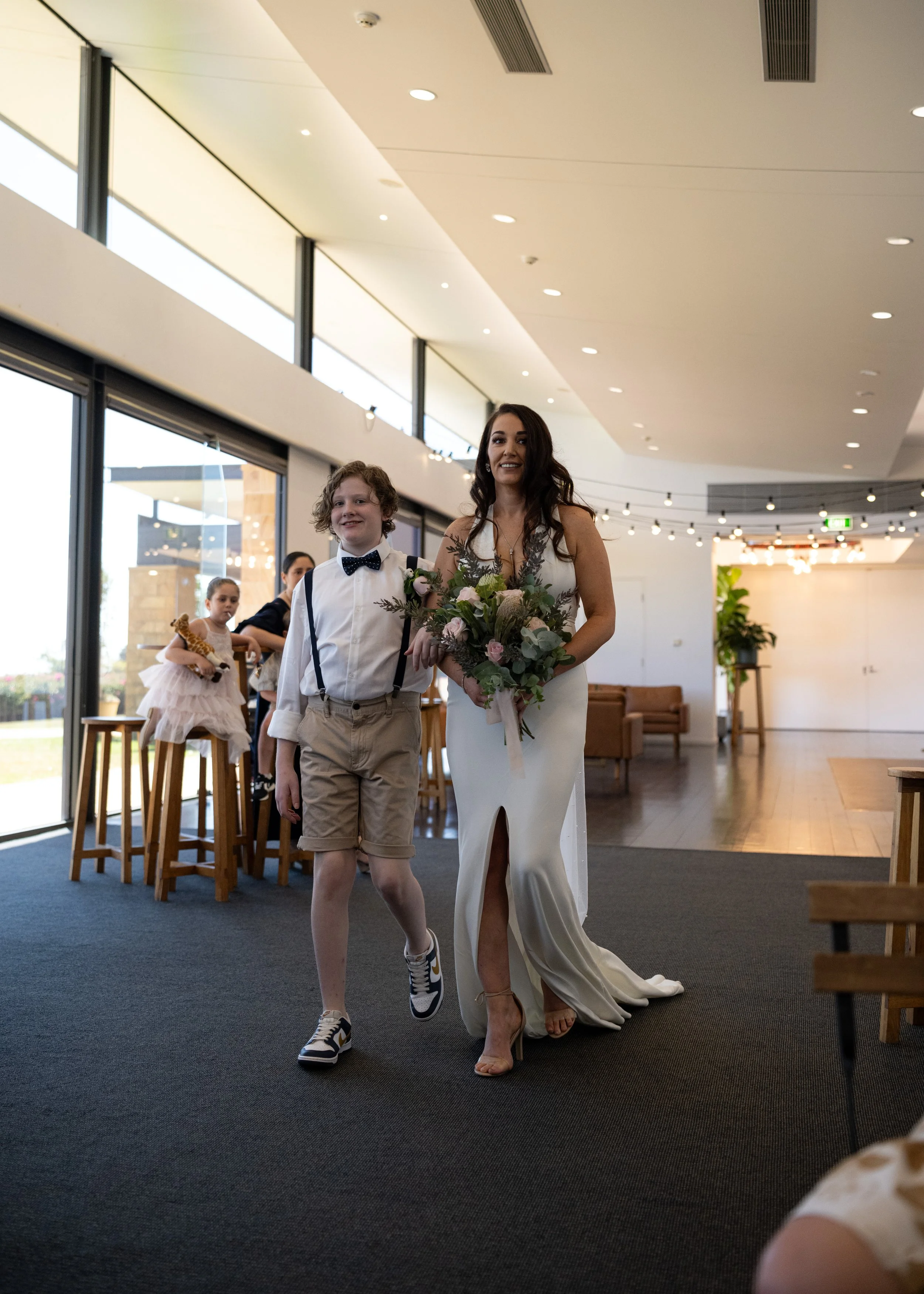 A woman in a white bridal gown holding a bouquet of flowers walking with a young boy in a white shirt, beige shorts, and a black bowtie inside a modern, well-lit building with large windows, other children sitting at a high table in the background.