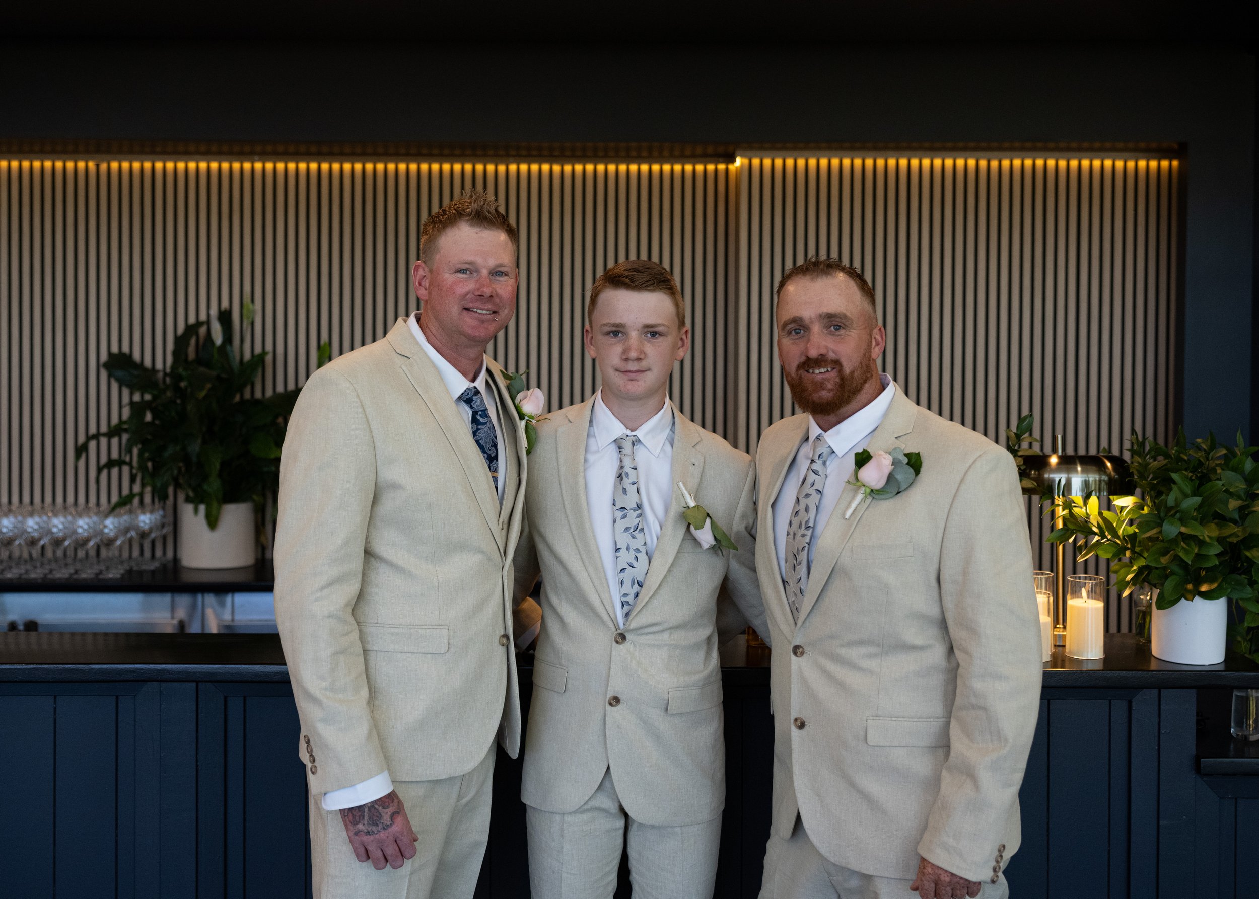 Three men dressed in cream-colored suits standing together indoors, smiling at the camera, with candles and potted plants in the background.